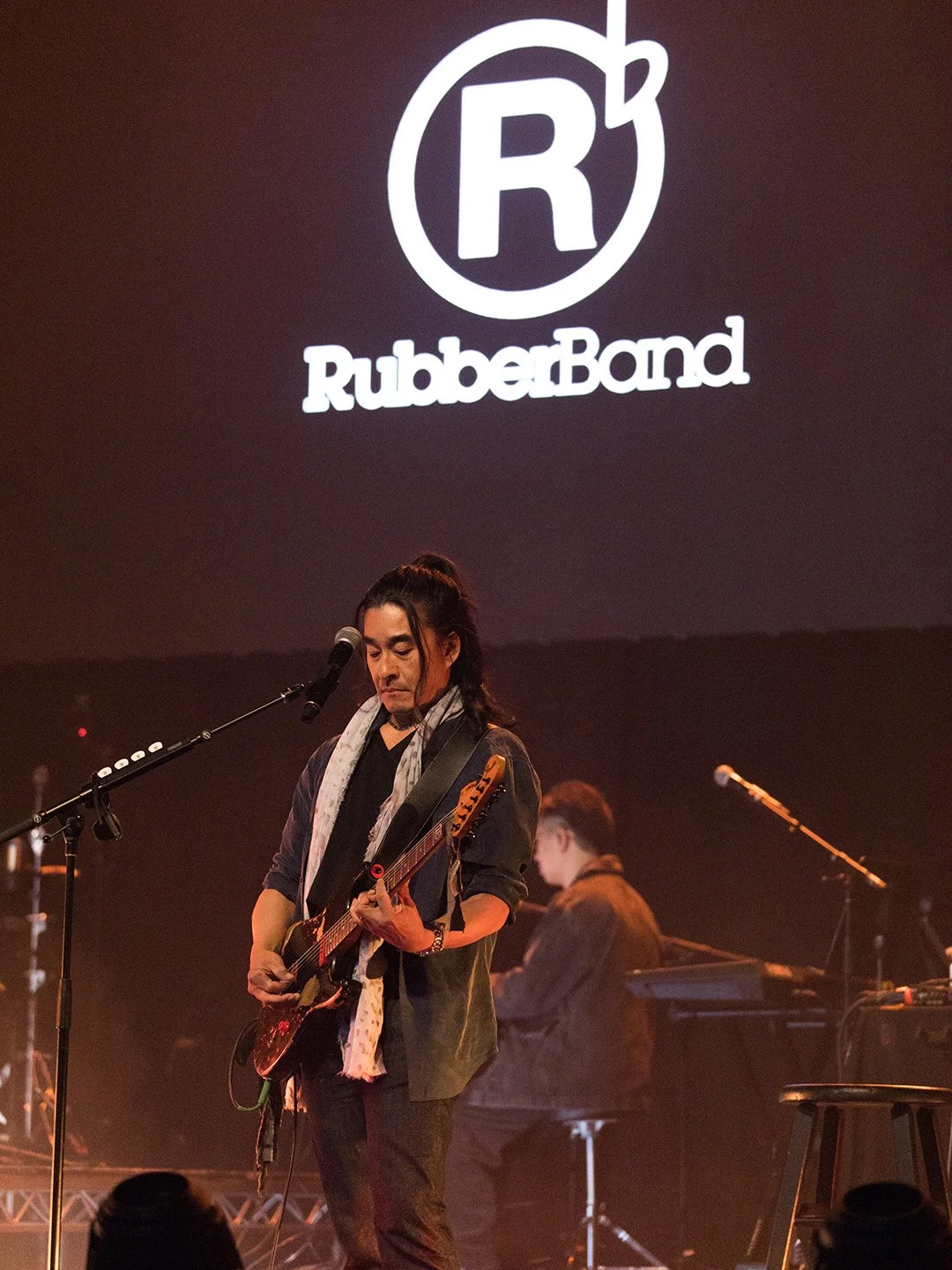 Female musician playing electric guitar on stage with a keyboard player in the background at a RubberBand concert, with the RubberBand logo projected on the screen above.