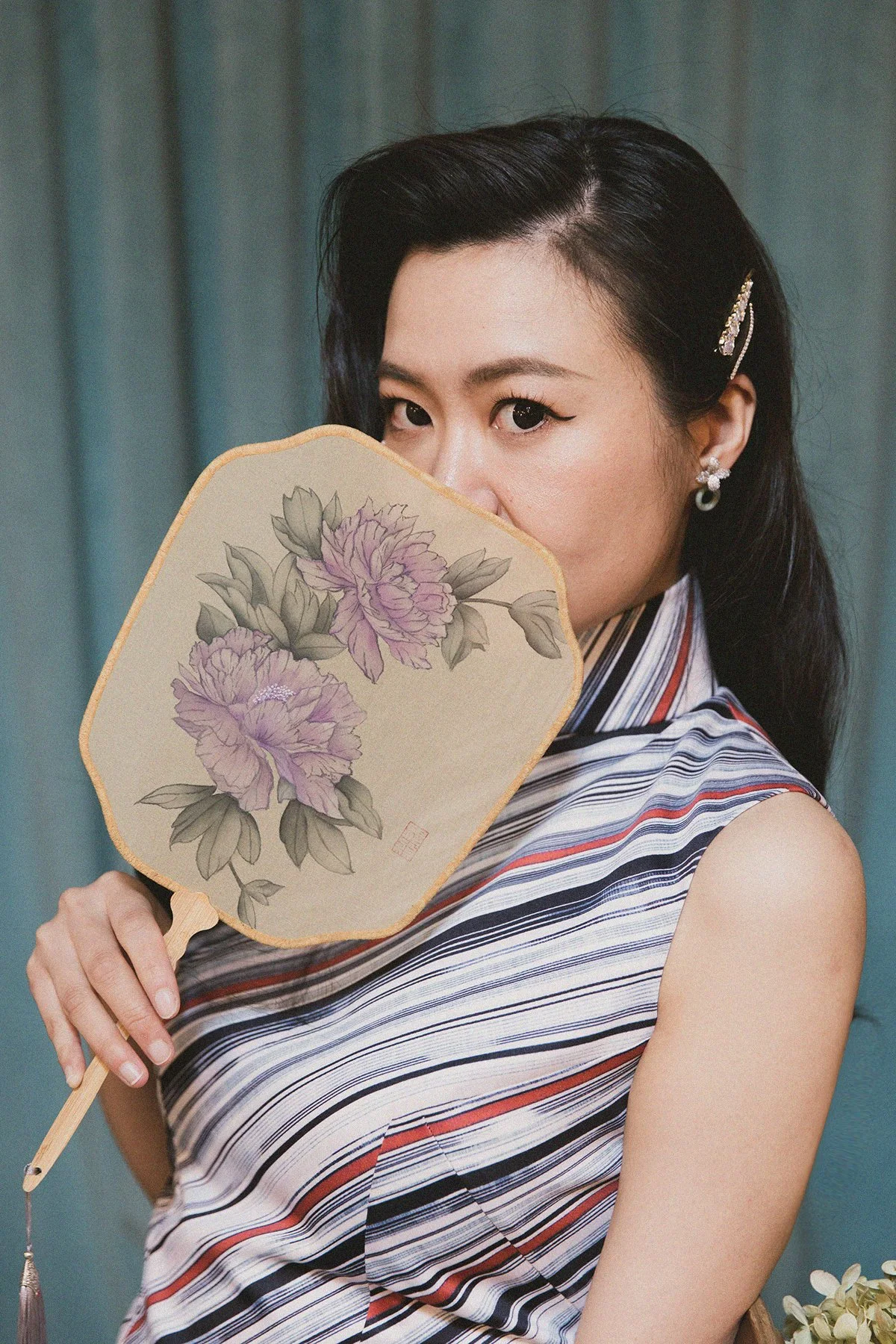 A woman with black hair and earrings holding a decorative fan with purple flowers in front of her face, partially covering her nose and mouth.