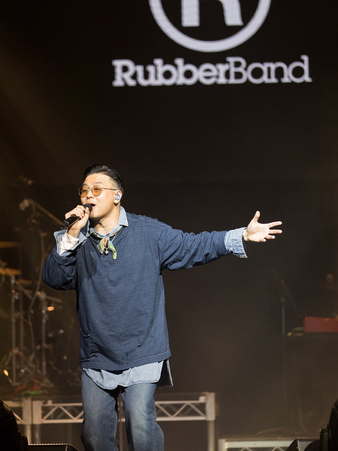 A male performer singing on stage with a microphone in hand, wearing glasses, a denim jacket over a shirt, and jeans, in front of a black backdrop with the RubberBond logo.