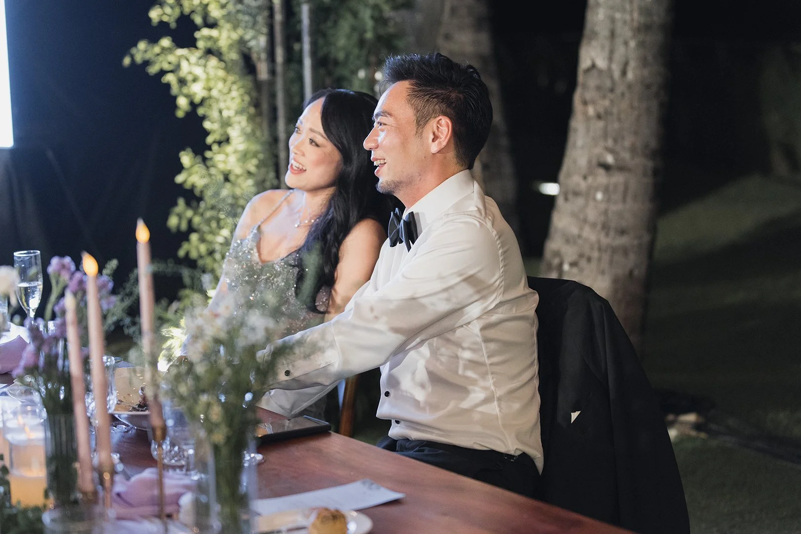 A happy couple, a woman with long black hair and a man with short black hair and a goatee, dressed in formal attire, sitting at a decorated outdoor event table with flowers and candles, smiling and engaging in conversation.