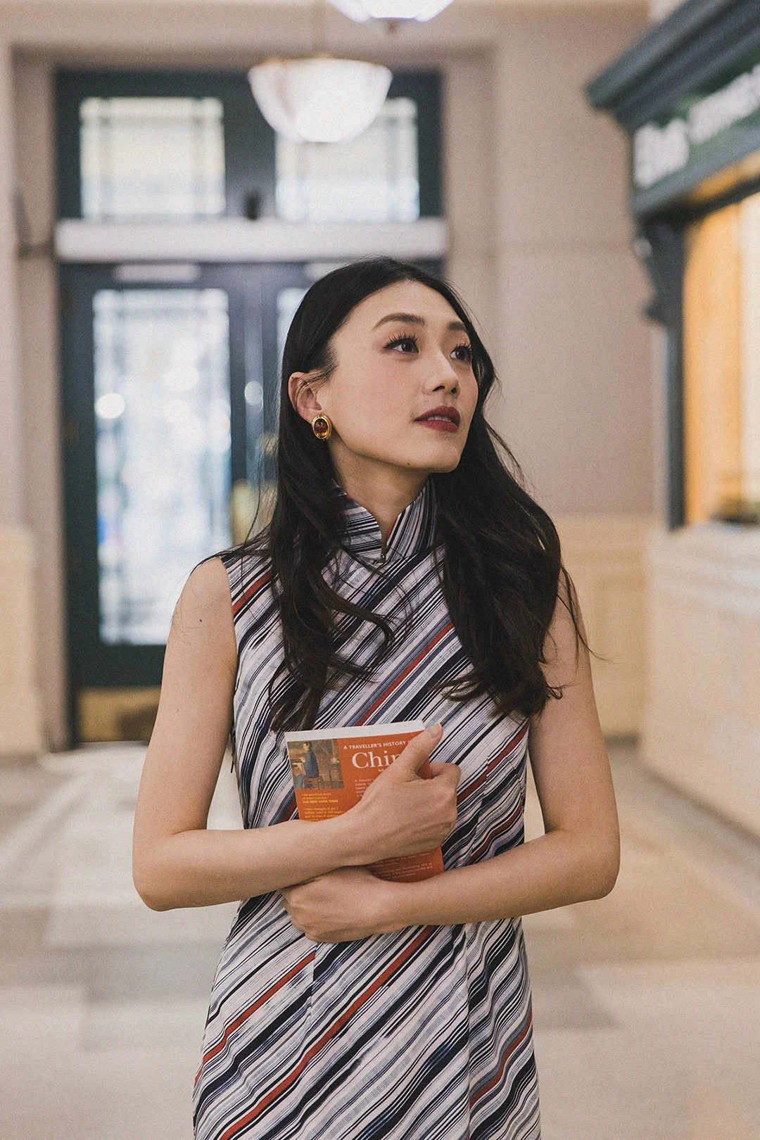 A woman with long dark hair, wearing gold earrings, holding a brochure or pamphlet, standing indoors near a door with glass panels and a notice board.