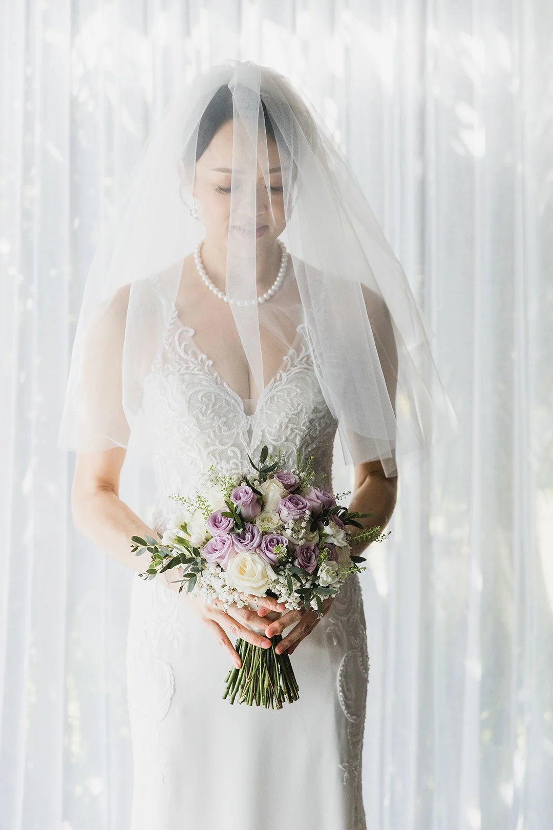 A bride standing in front of a sheer curtain, wearing a lace wedding dress, pearl necklace, and veil, holding a bouquet of lavender and white roses.