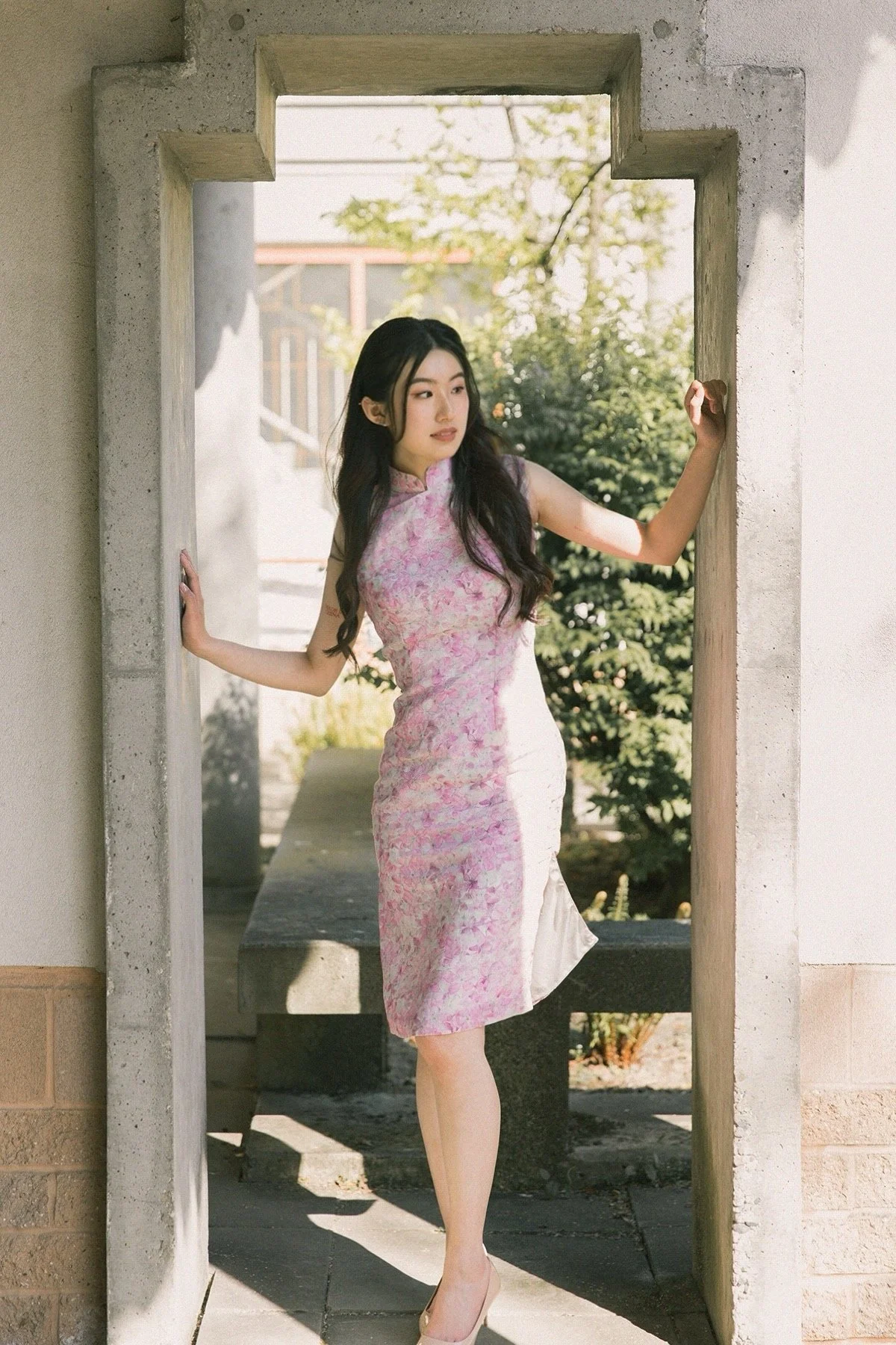 A young woman in a pink and white floral cheongsam dress stands in a stone doorway, with one hand on each side of the frame, looking to her right. Sunlight filters through nearby trees, creating a soft, natural light.