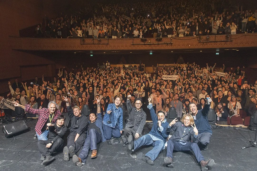 Group of performers on stage taking a selfie with a large, cheering audience in a theater or concert hall.