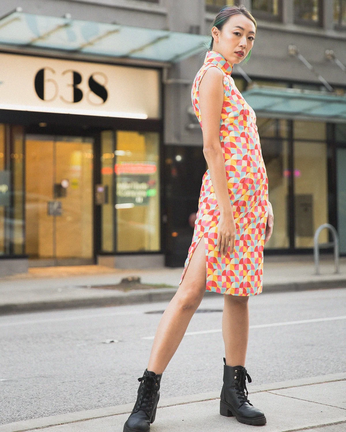 A woman standing on a city sidewalk in front of a building with the sign 'GBS'. She is wearing a colorful sleeveless dress with a high collar and side slit, paired with black combat boots. Her hair is tied back and she is looking at the camera.