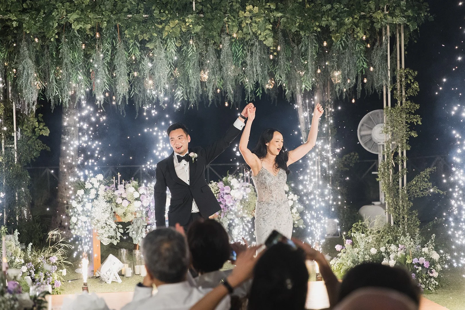 A newlywed couple dances and celebrates at their wedding reception, surrounded by wedding decorations, flowers, and sparkling lights.