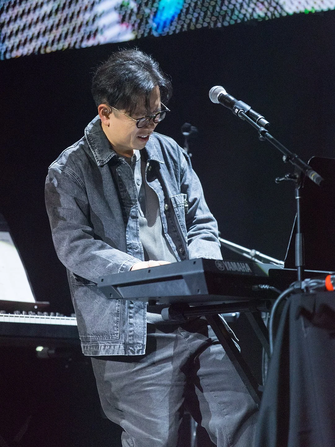 Man playing Yamaha keyboard, singing into a microphone on stage, wearing glasses and a denim jacket.