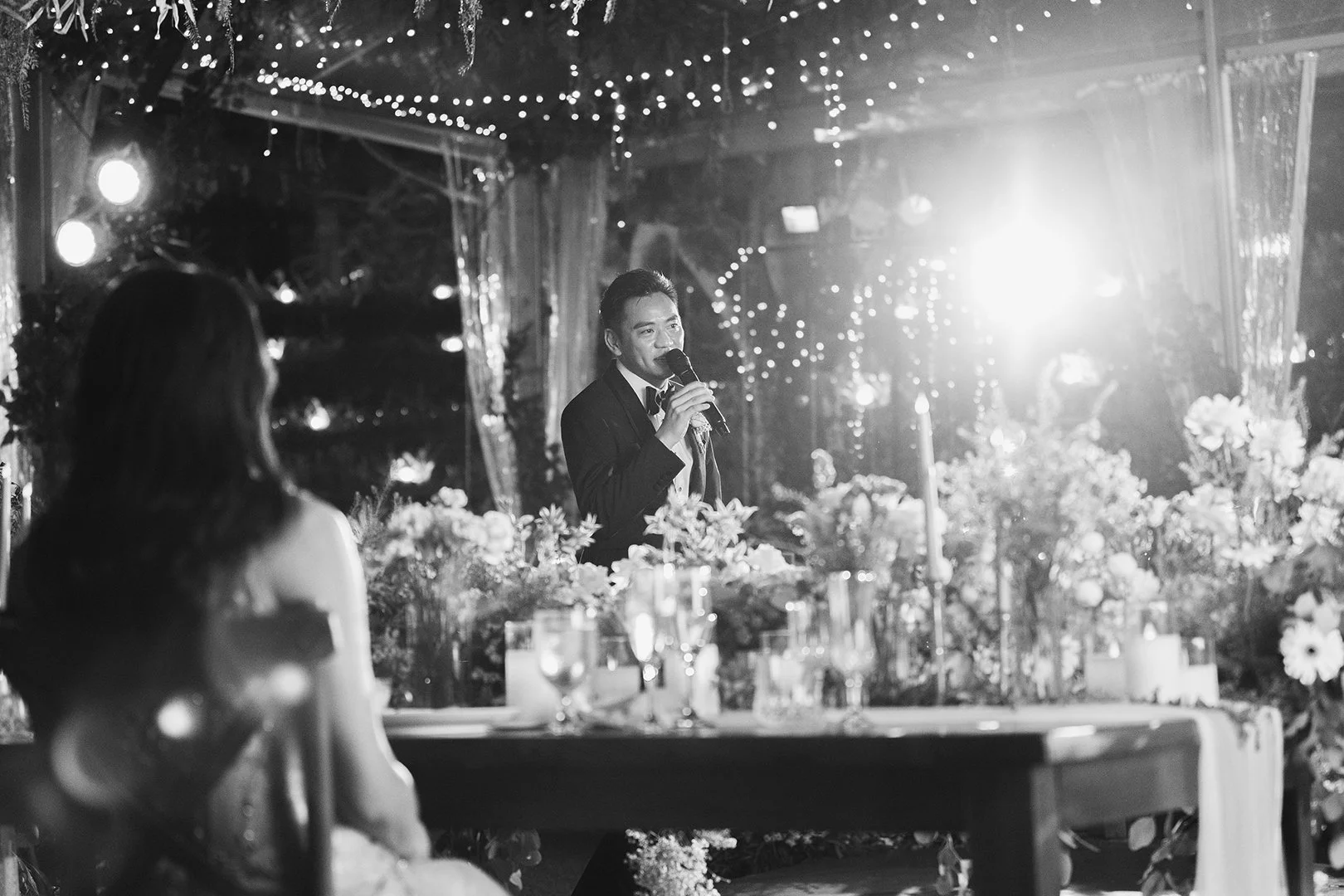A man in a tuxedo speaking into a microphone at a wedding reception decorated with flowers and string lights, with a woman in a dress sitting at the table in the foreground.