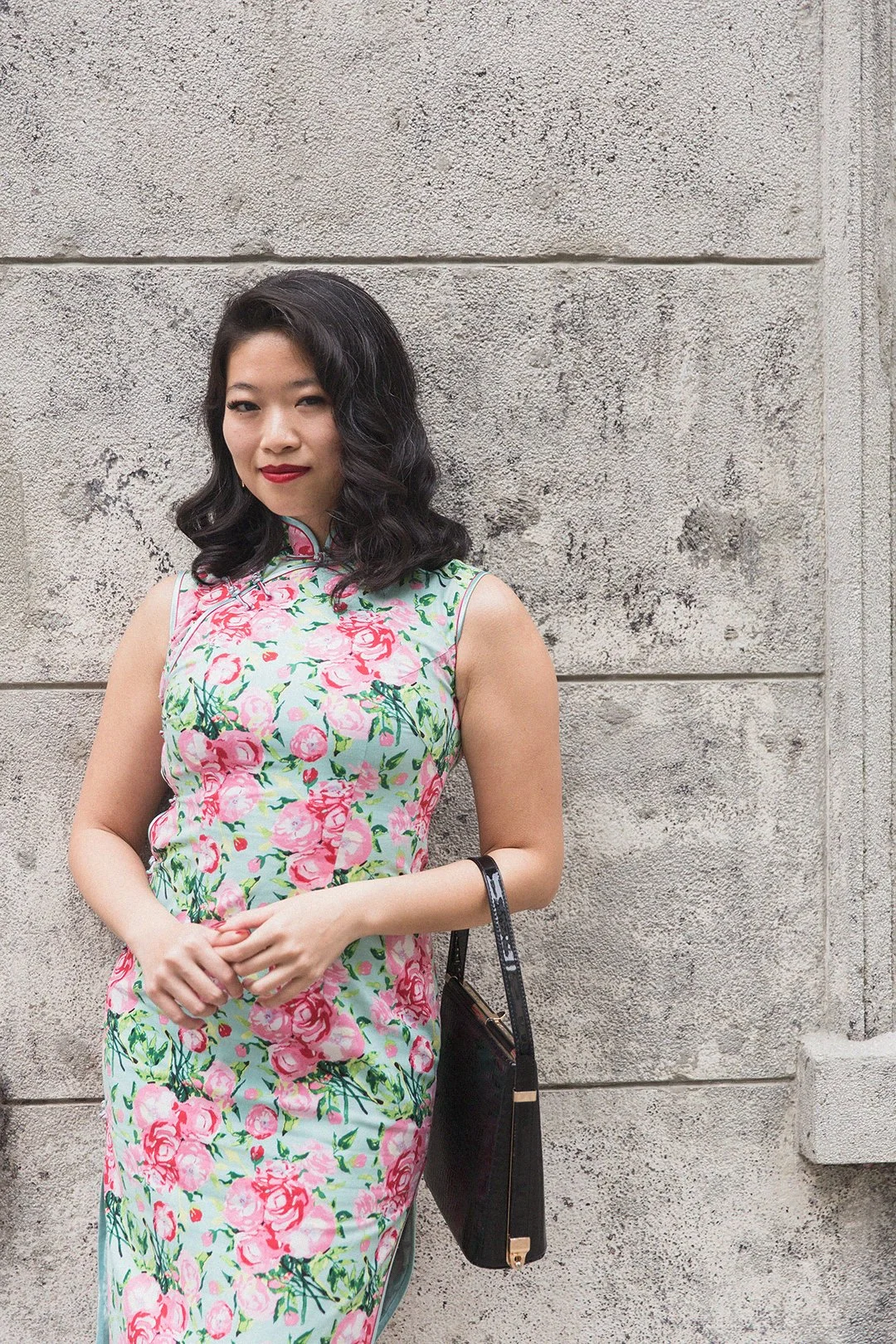 A woman with black, wavy hair and red lipstick wearing a sleeveless floral cheongsam dress, standing against a textured gray wall, holding a black handbag.