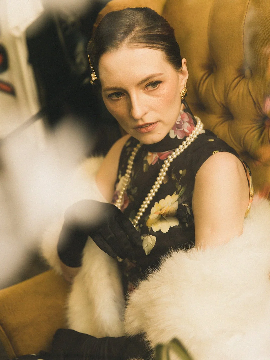 A woman with dark hair, wearing a floral dress, pearl necklace, and gold jewelry, sitting on an orange tufted armchair with a fur throw, looking slightly to her right.