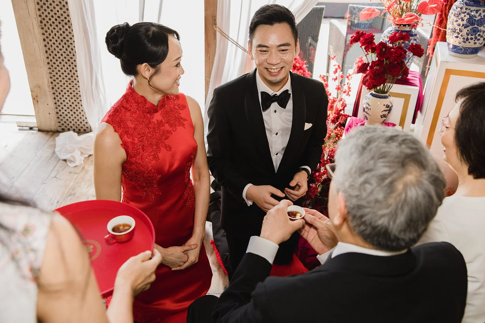 A traditional Chinese tea ceremony with a woman in a red dress and a man in a tuxedo, surrounded by family members in a decorated room with red flowers.