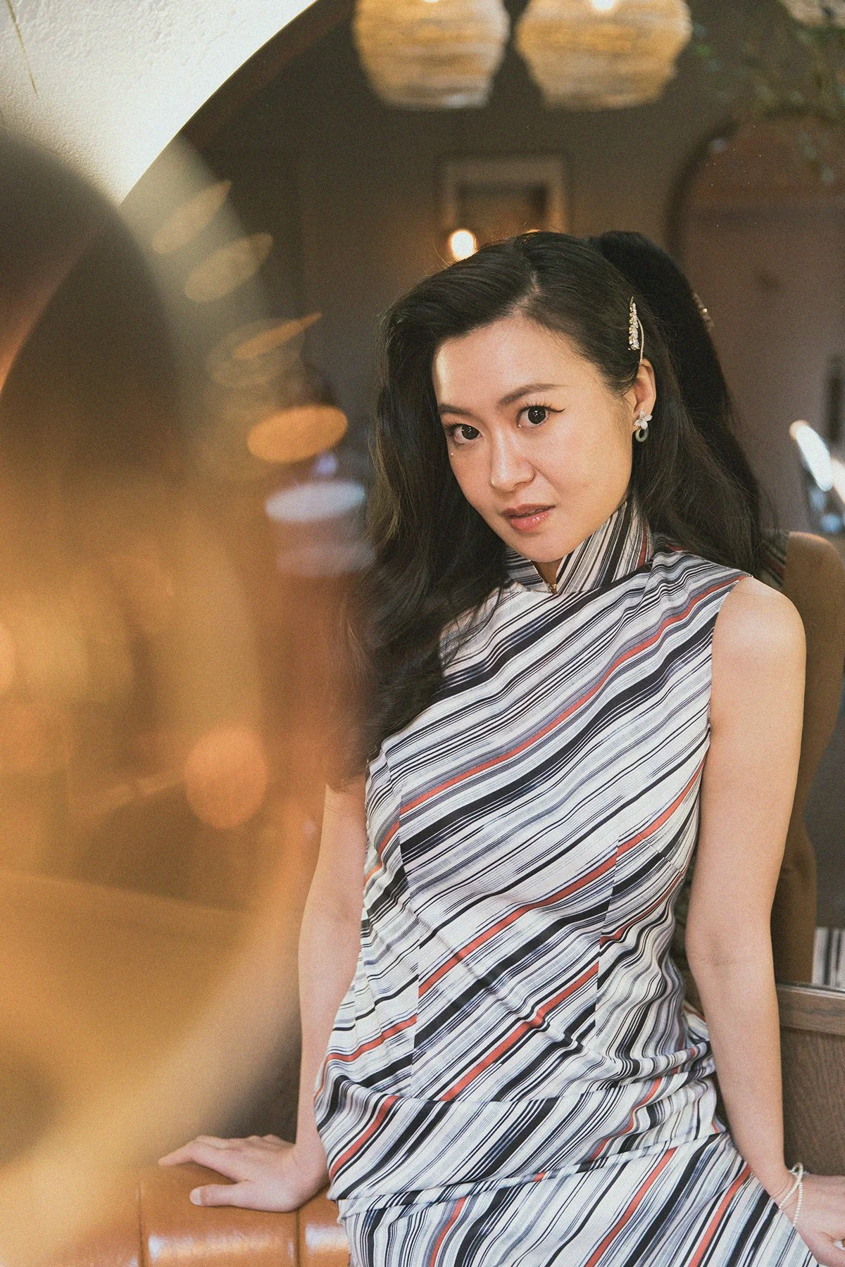A woman with long black hair wearing a sleeveless striped dress sitting at a restaurant or cafe, looking at the camera.