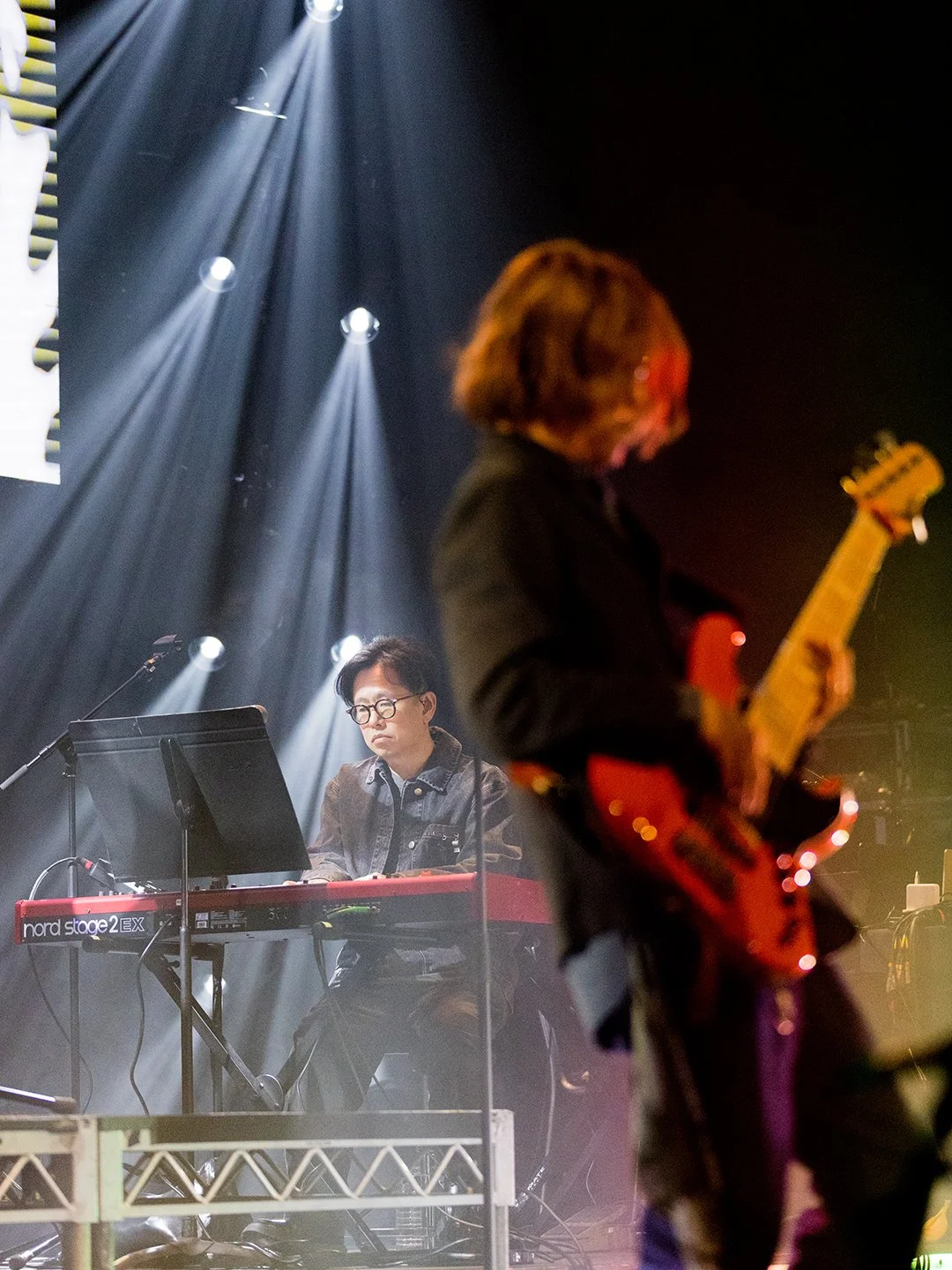 Musicians performing on stage with lights shining down. One person playing keyboard and another person playing guitar with their face and upper body visible.