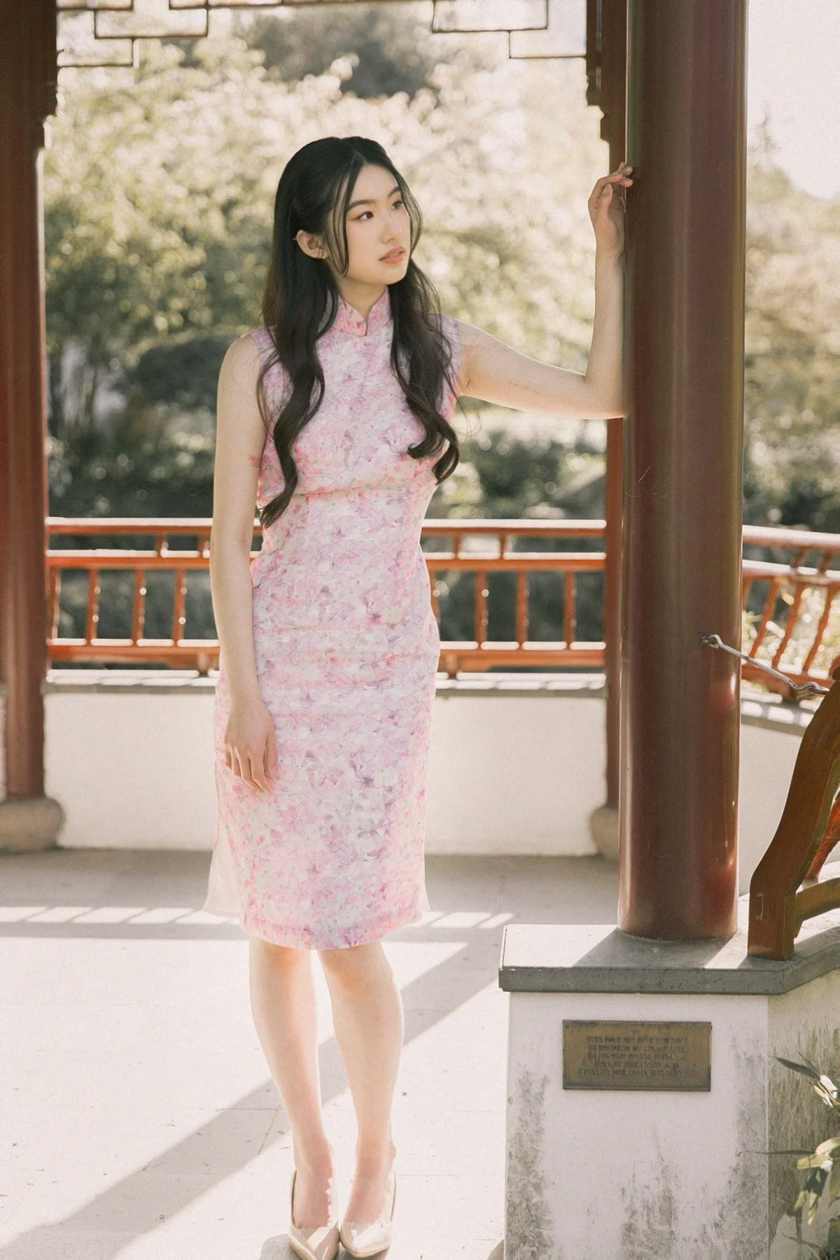 A young woman in a pink floral qipao dress standing on a balcony, holding onto a wooden pillar, with trees and a traditional railing in the background, during daytime.