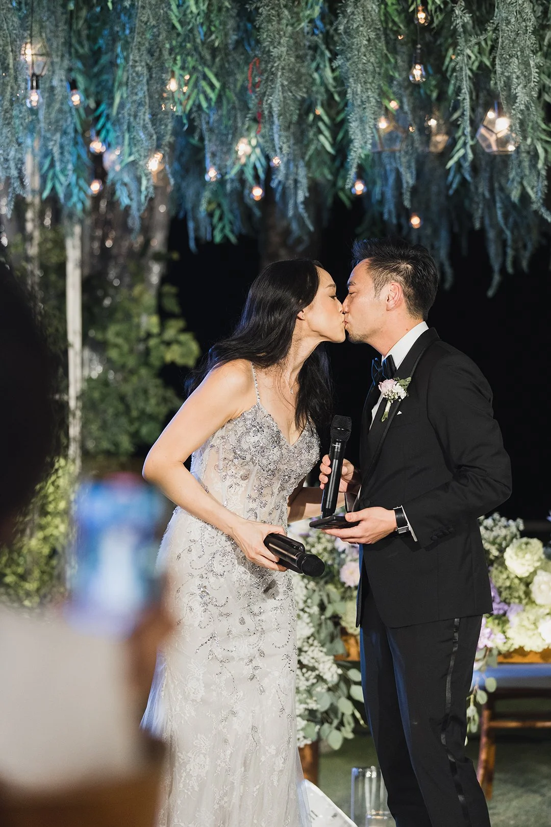 A couple in wedding attire kissing under a decorated canopy with hanging greenery and string lights, at night.