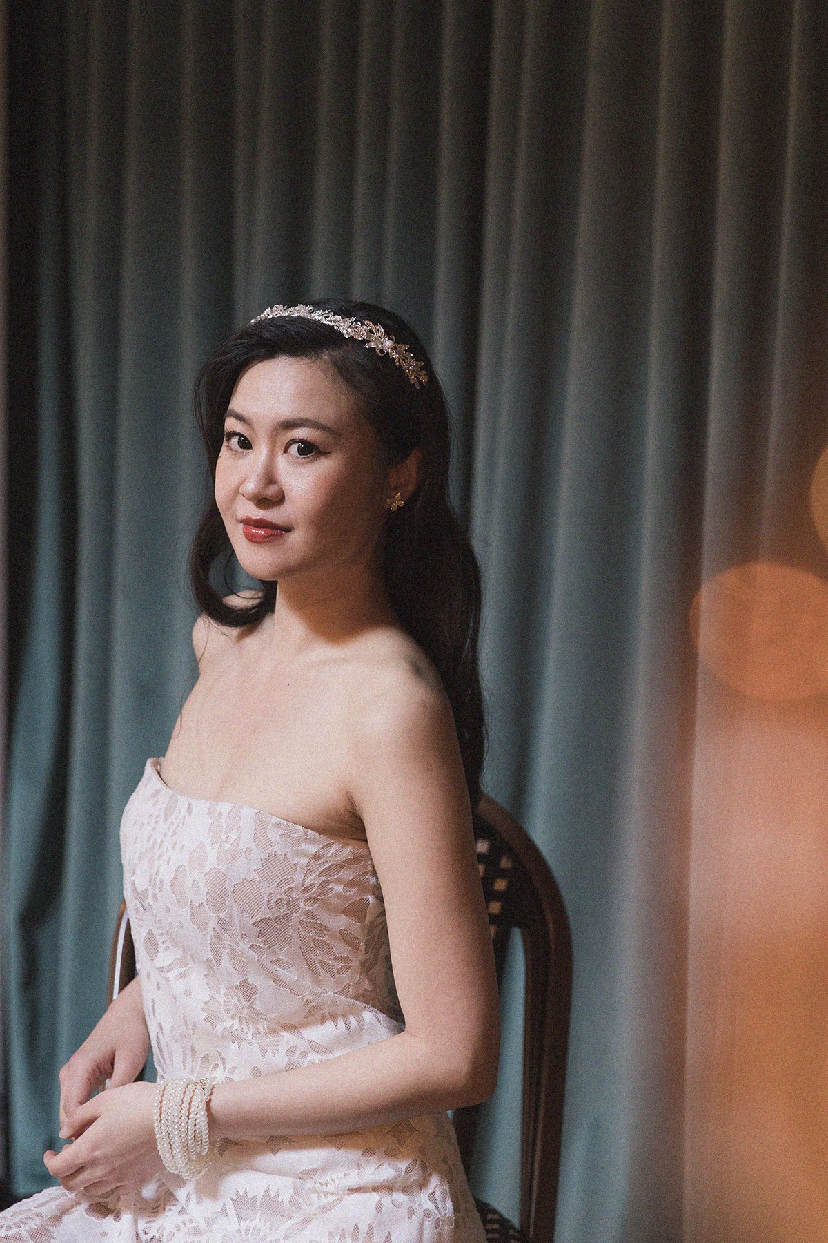 A young woman with dark hair wearing a strapless lace dress and a tiara, sitting on a chair in front of green curtains.