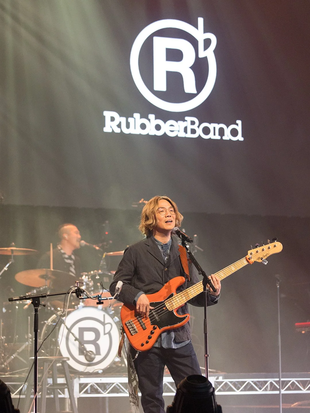 Musician with shoulder-length hair and glasses playing an orange electric bass guitar on stage, with a drummer in the background and a large screen displaying the RubberBand logo.