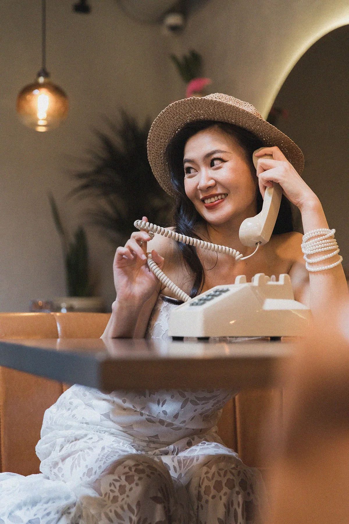 A woman wearing a wide-brimmed hat and white lace outfit, sitting at a table, talking on a beige rotary phone, smiling, with a background of a potted plant, warm-toned lighting, and a hanging light fixture.