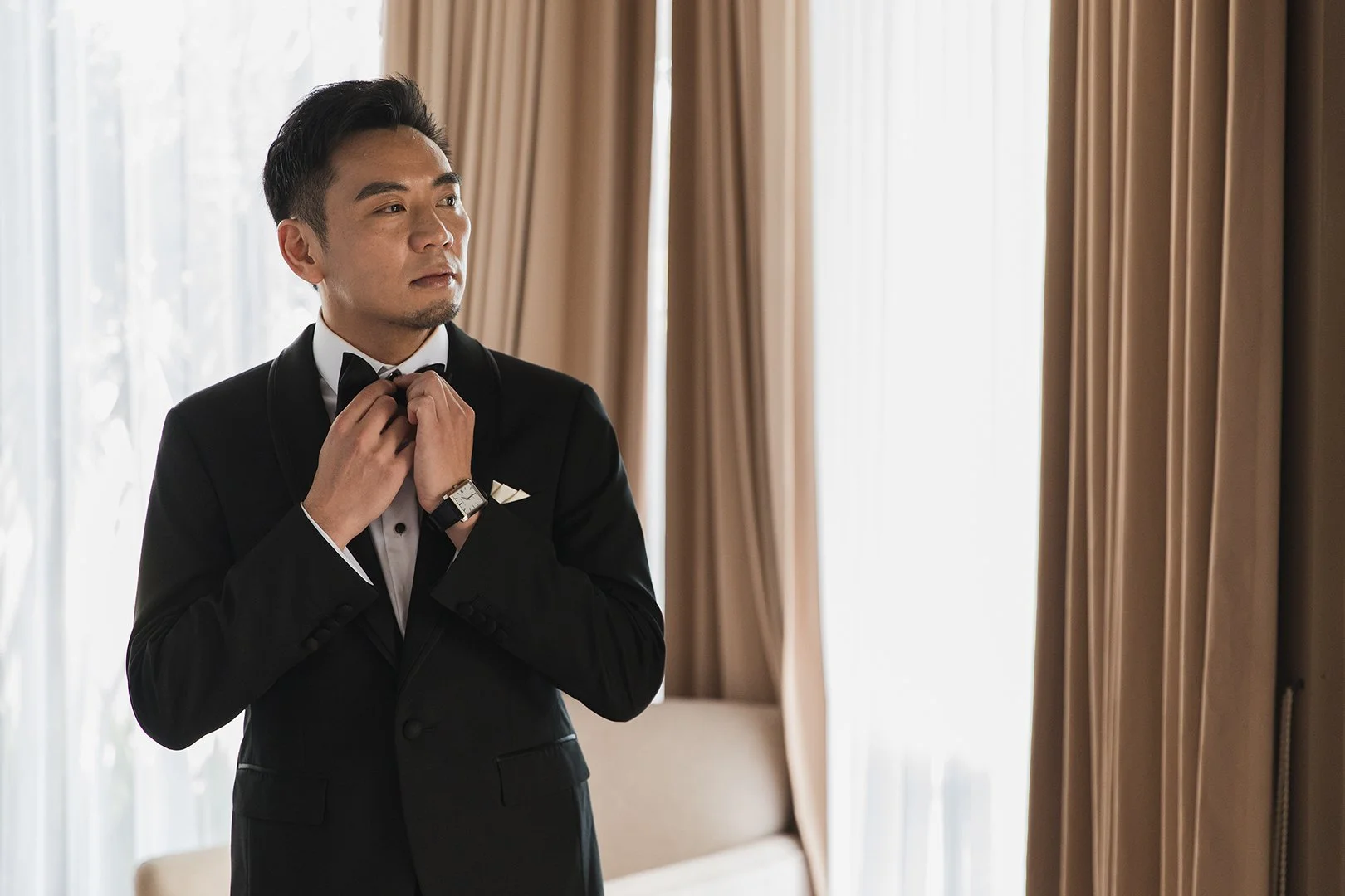 A man in a black tuxedo adjusting his bow tie in a well-lit room with large windows and beige curtains.