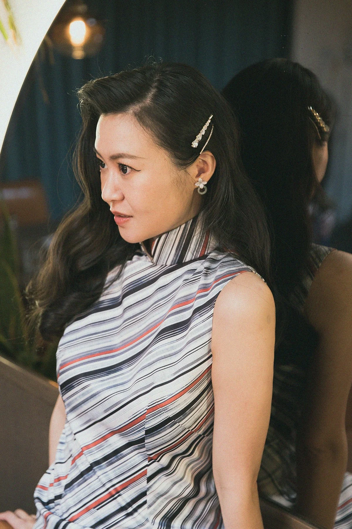 A woman with long dark hair, wearing a sleeveless striped top and decorative hair clips and earrings, looking to her left in front of a mirror.