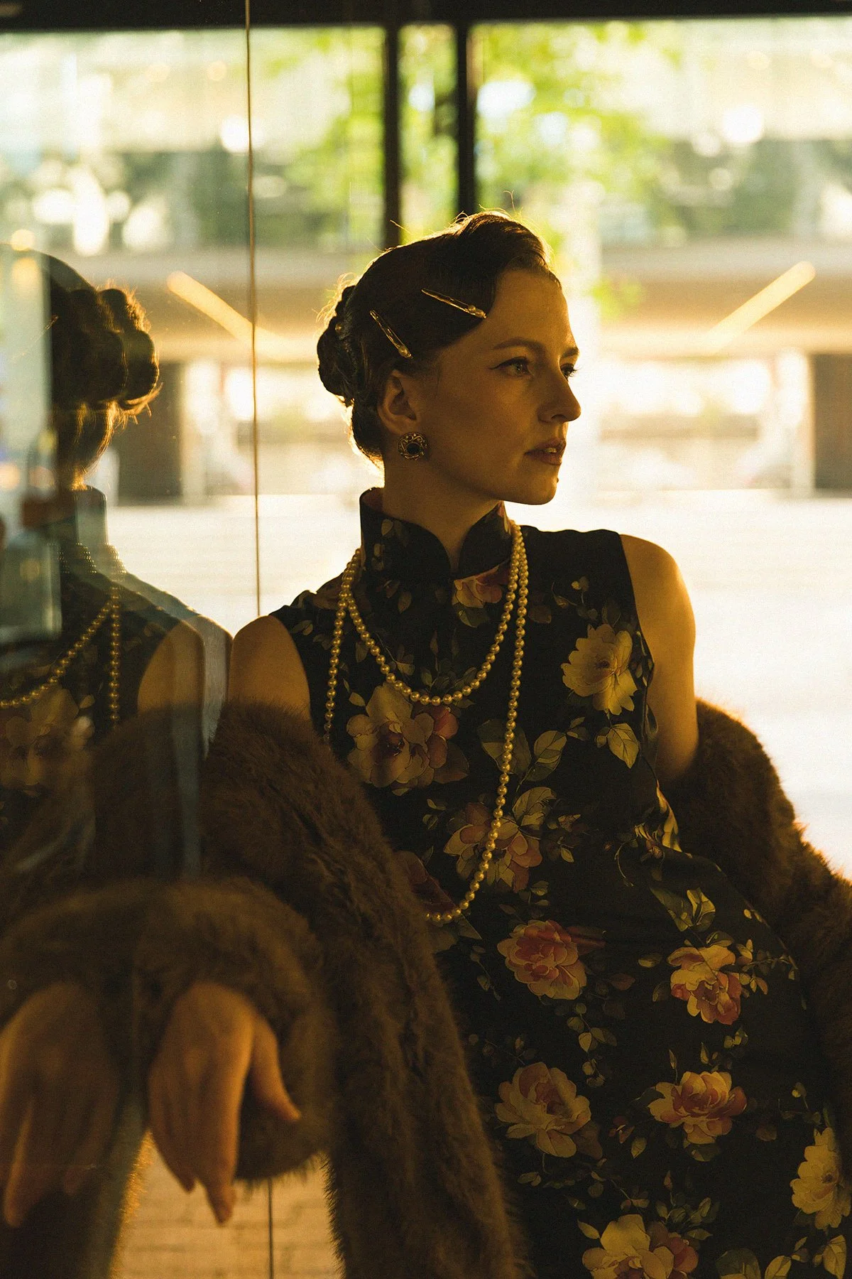 A woman with vintage hairstyle and pearl jewelry sitting inside a dimly lit space, reflected in a glass wall with a city street in the background.