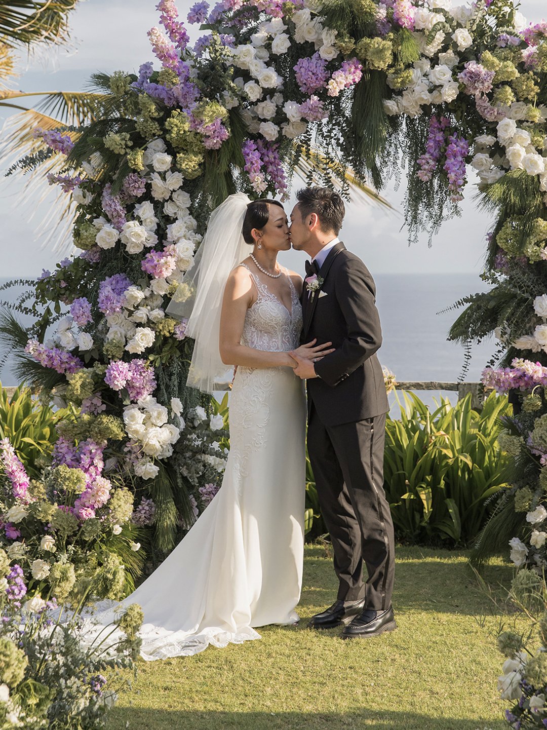 A bride and groom sharing a kiss during their outdoor wedding ceremony beneath a floral arch with purple, white, and green flowers, overlooking the ocean.