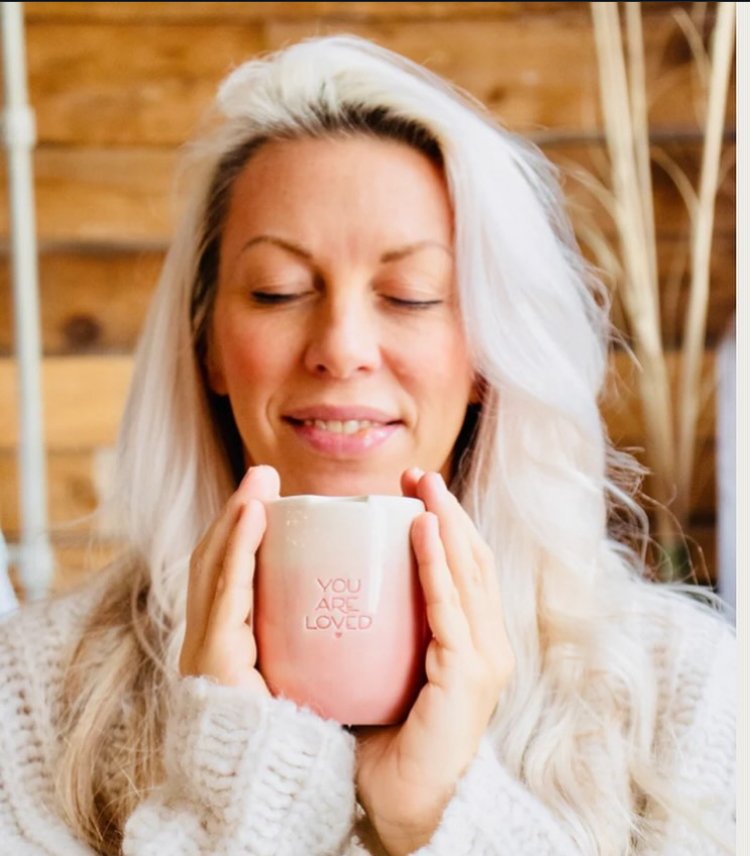 Woman with long blonde hair holding a pink mug with the words "You Are Loved" written on it, smiling and with eyes closed.