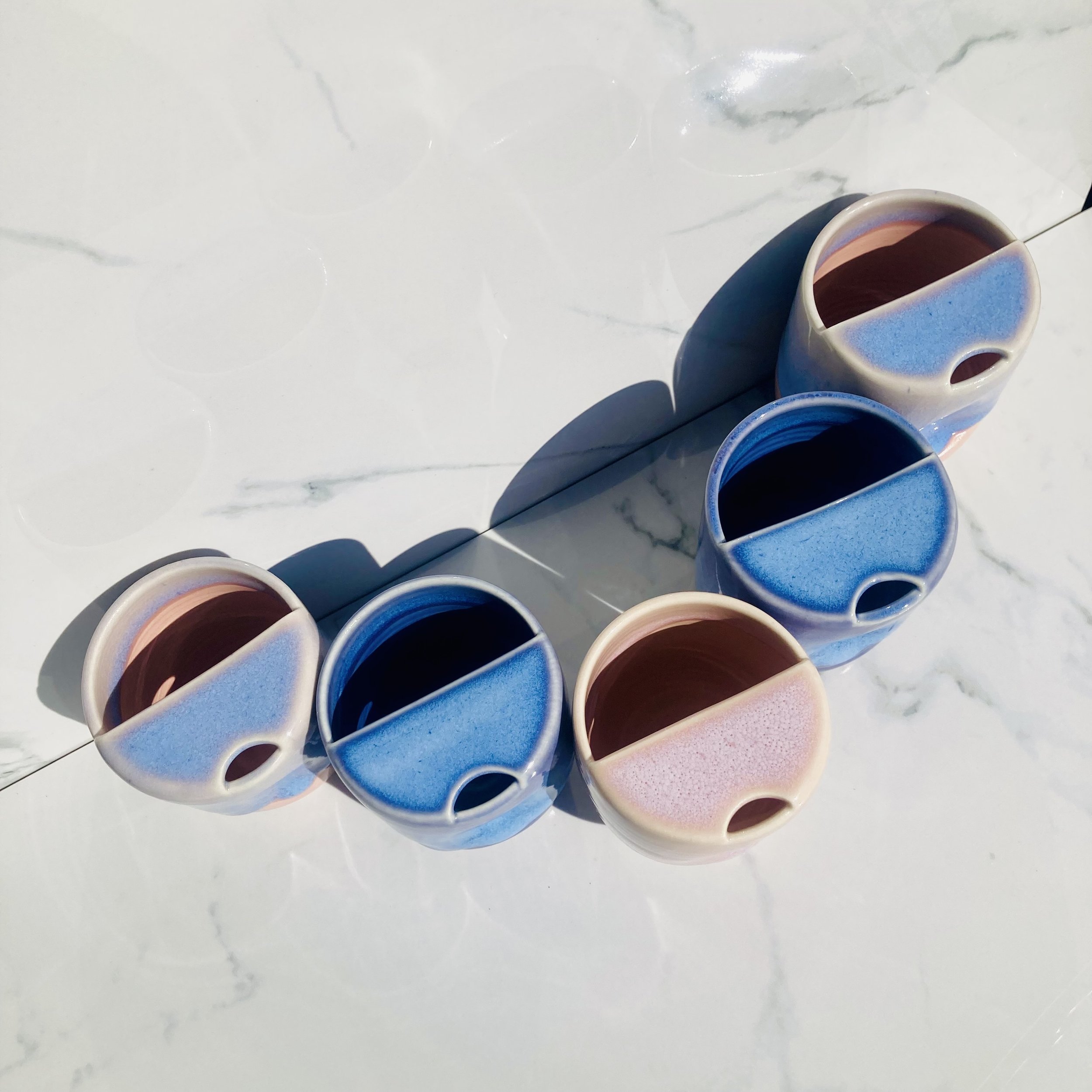 Five colorful ceramic cups filled with dark liquid, arranged on a white marble surface with shadows cast by sunlight.