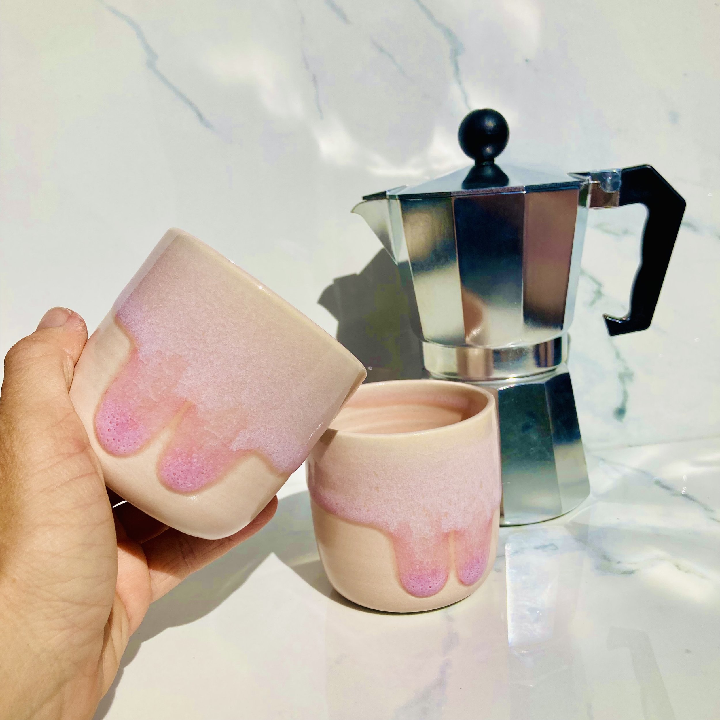 Two pink ceramic cups with a drip glaze design, a hand holding one cup, and a silver moka pot on a white marble surface.