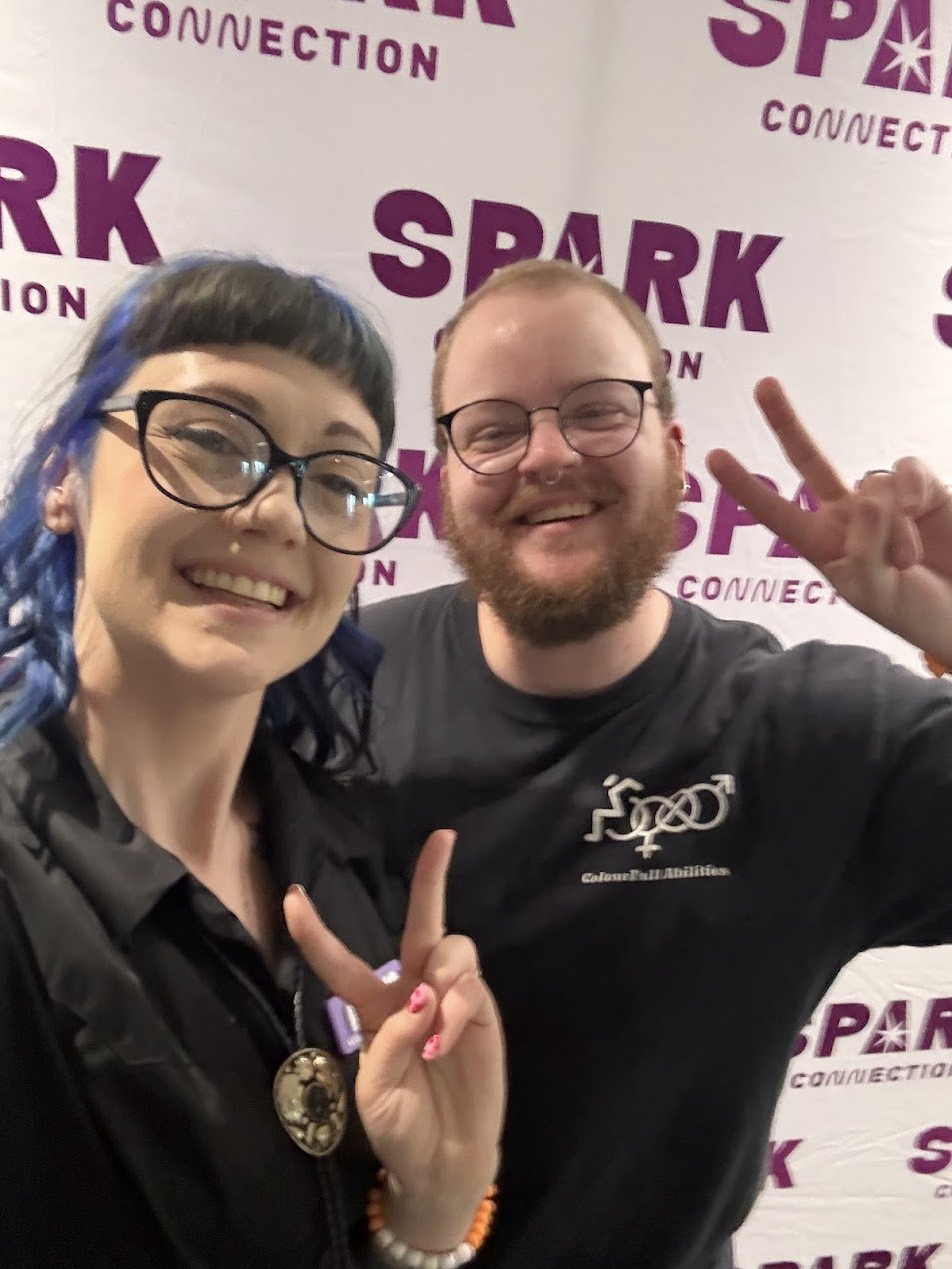 Amelia is pictured smiling in black dress and glasses, holding a peace sign with another person who is smiling and wearing a colourfull abilities shirt, the banner behind them says "spark connection"