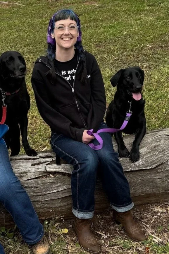 Amelia is sitting on a log in jeans and a jumper, with her black labrador, they are smiling at the camera while her dog is has her tongue out