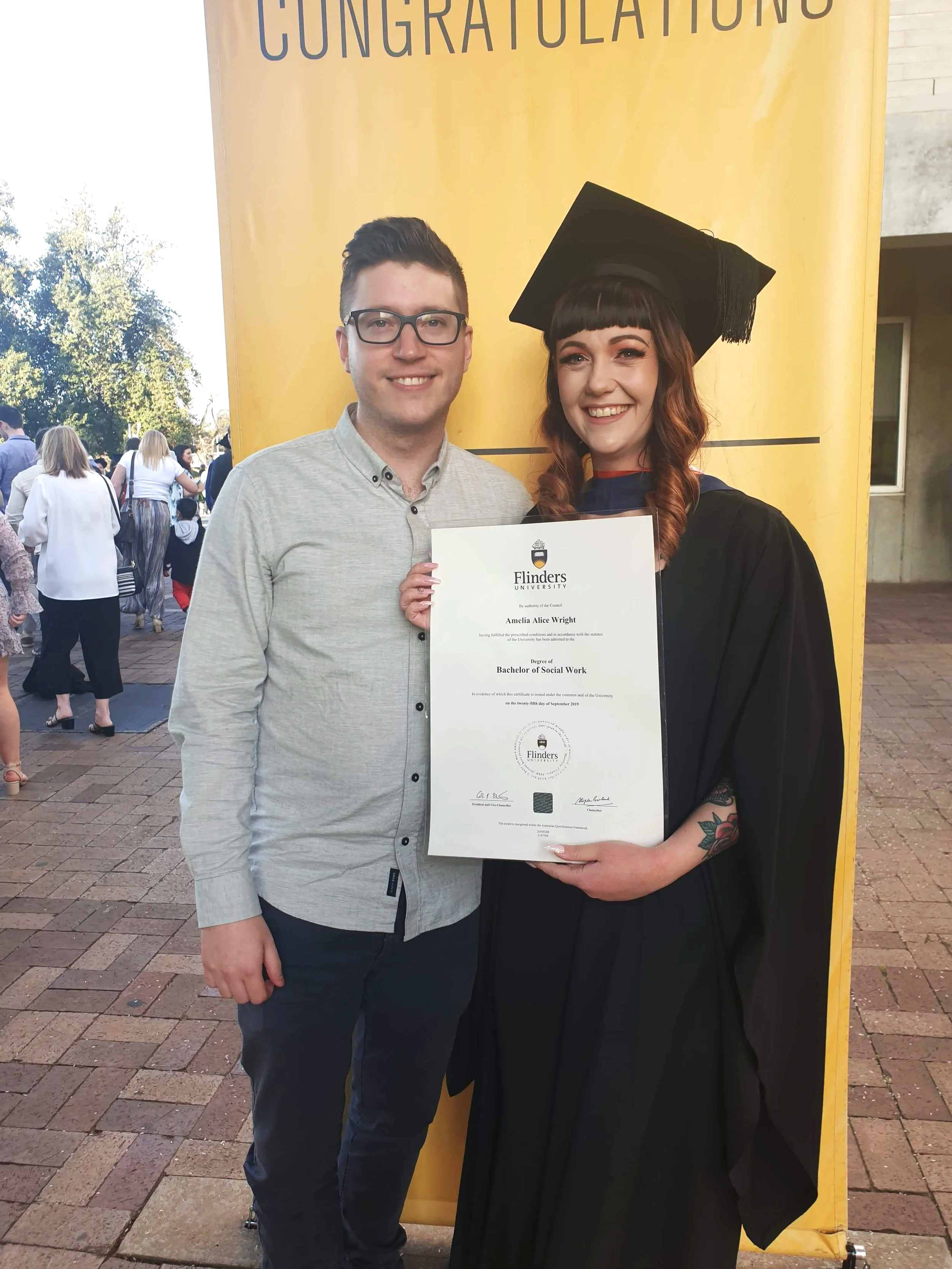 Amelia is wearing graduation attire, holding her bachelor of social work degree, she is dressed up with make up and her hair is curly, she is standing with a man (who is her partner Tom) who is wearing a light button up shirt, they are smiling