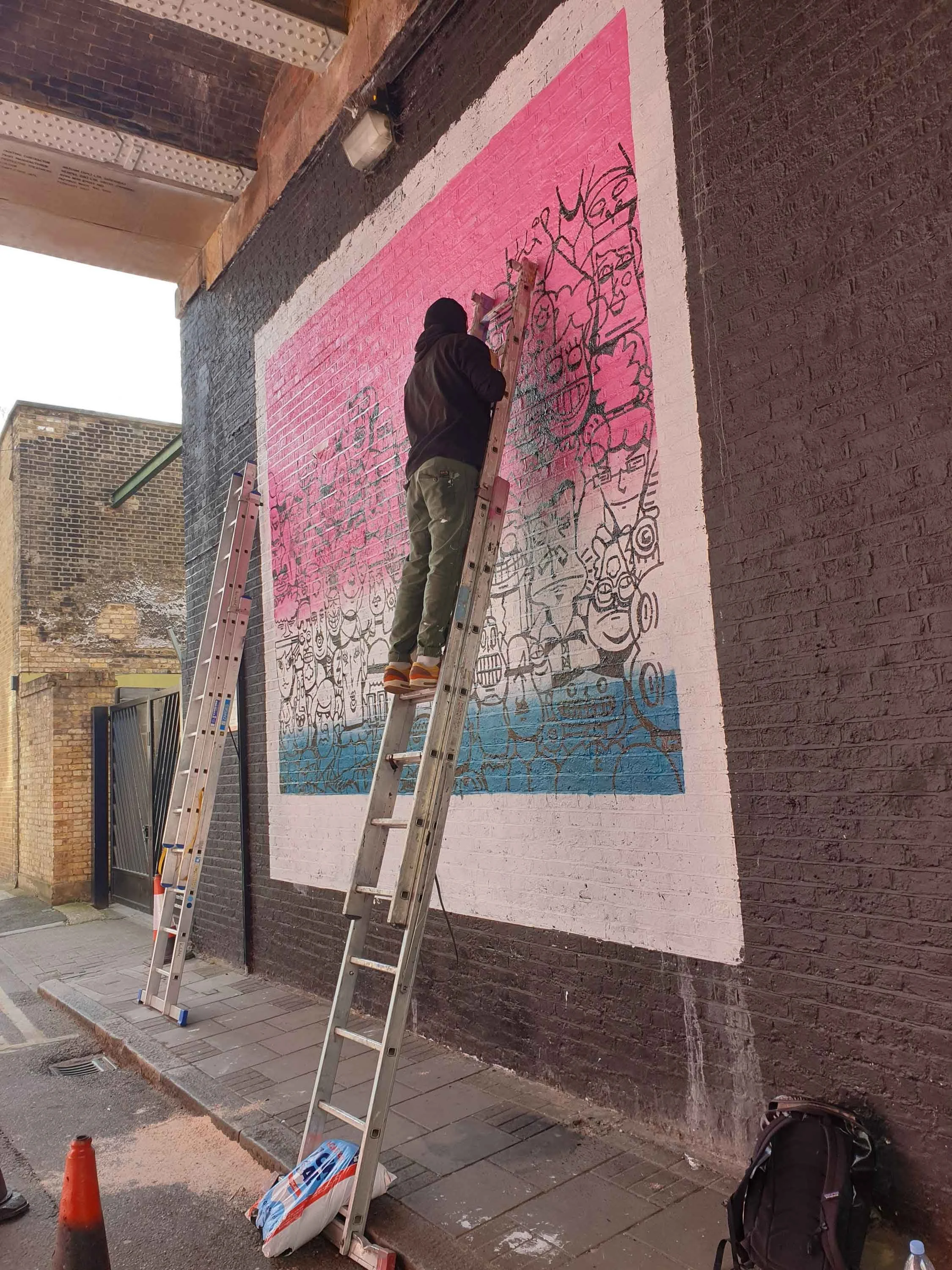 Chris Martin painting a large-scale street mural in Shoreditch London – in-progress photograph of artist on ladder applying black ink character illustrations over pink and teal painted wall under railway arch