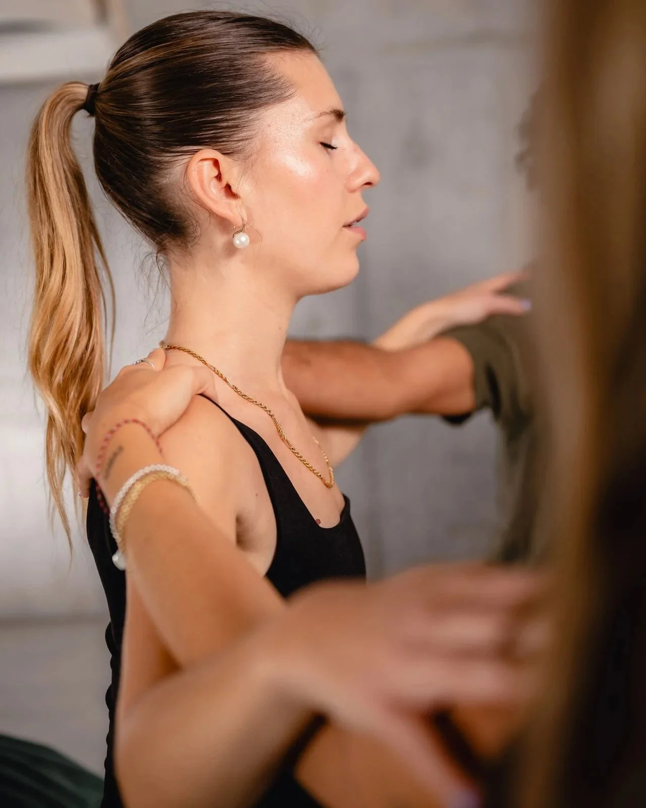 A woman with her eyes closed, wearing a black tank top, gold necklace, earrings, and bracelets, with her hand resting on her shoulder while someone else touches her arm.