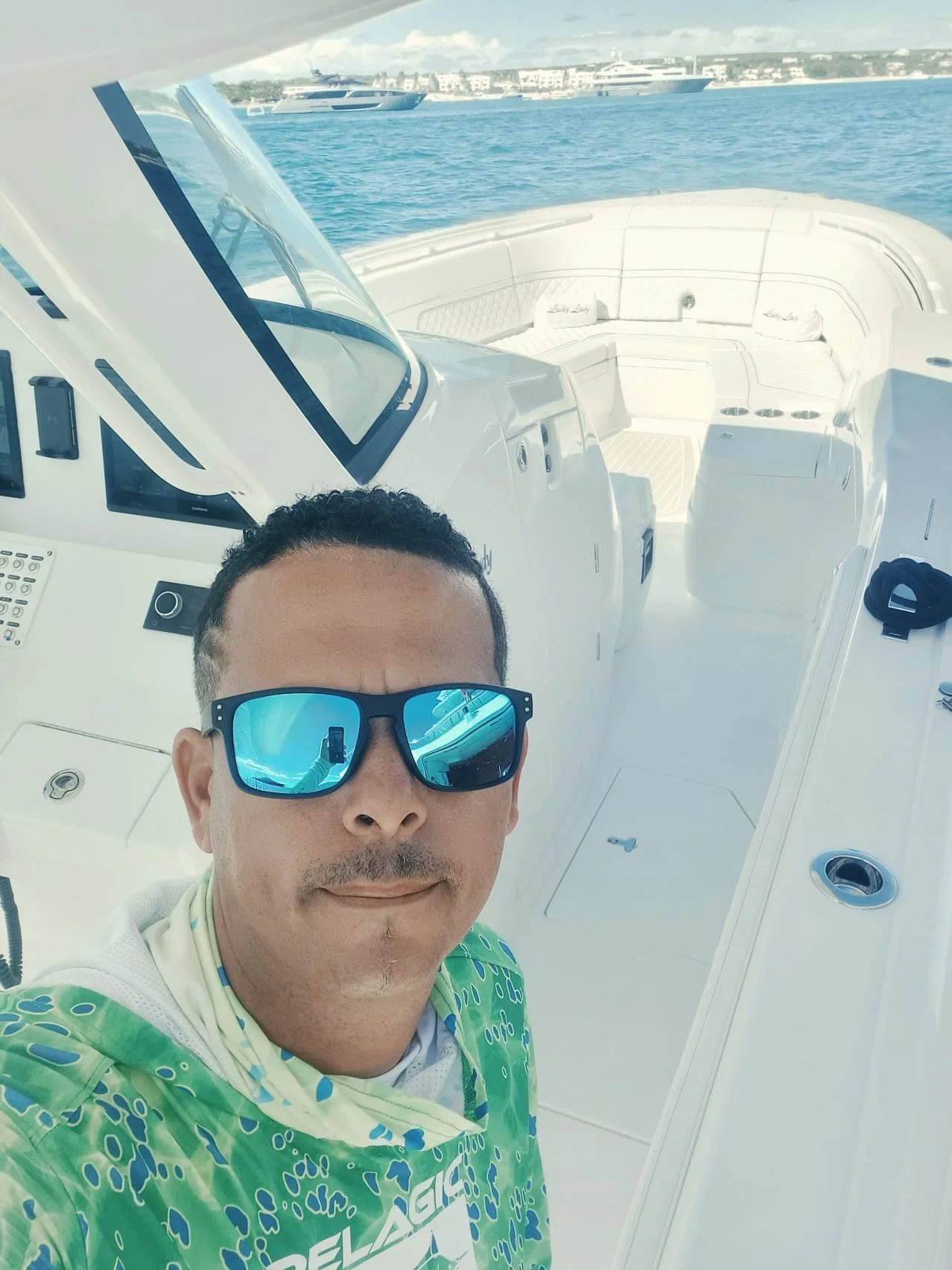 A man taking a selfie on a white boat or yacht, with water and other boats visible in the background. He is wearing blue sunglasses and a colorful green and white shirt.