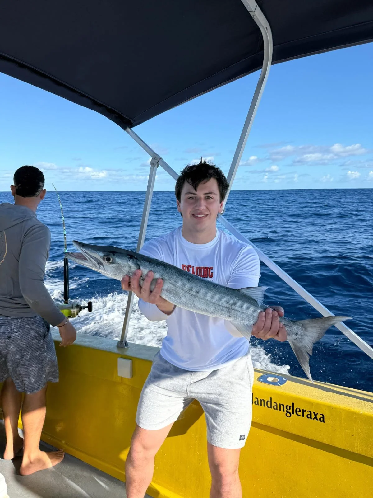 A young man on a boat holding a large fish, with another person fishing in the background, the ocean and sky visible.