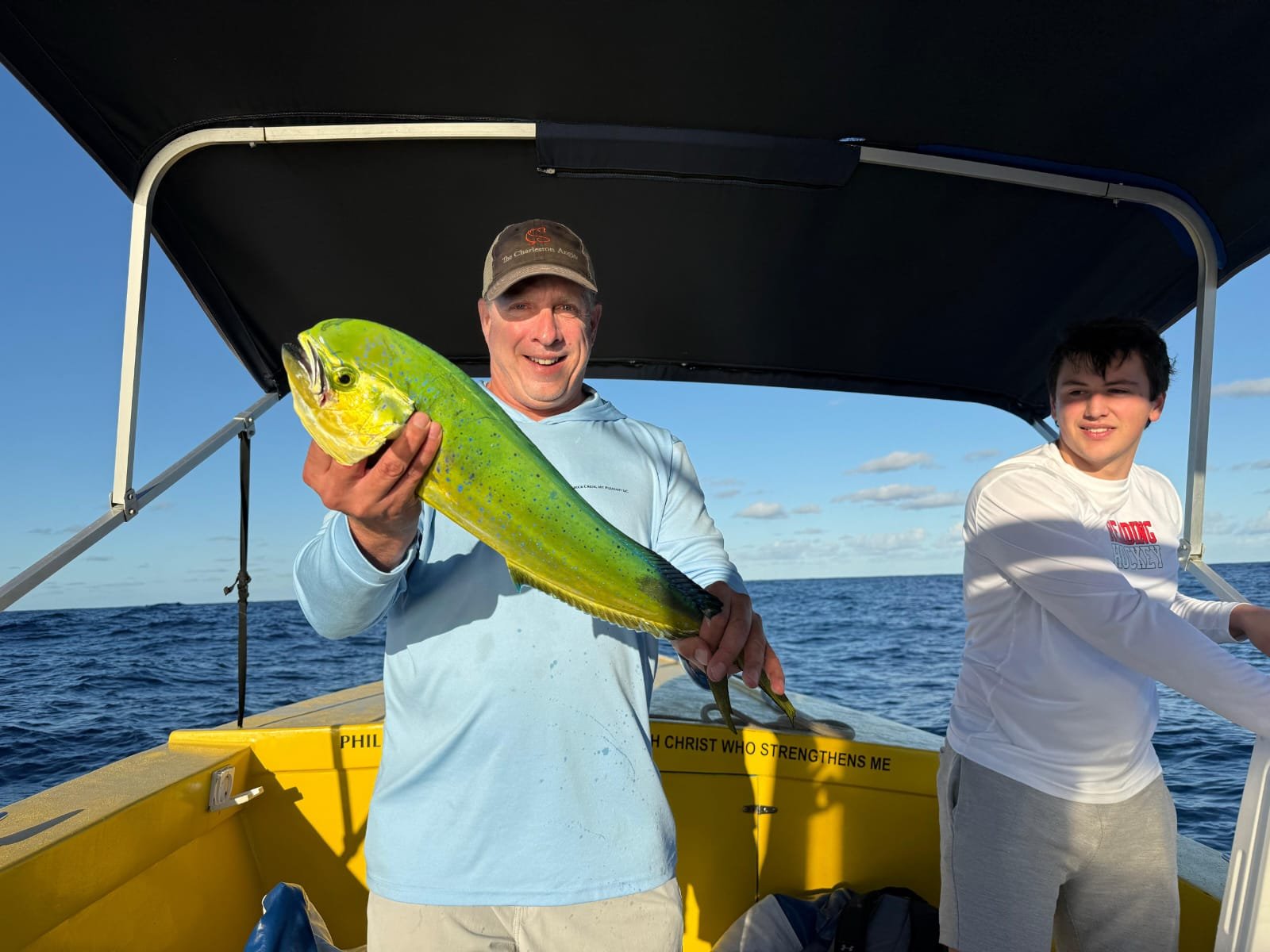A man holding a large, colorful fish on a boat with two people, ocean, and blue sky in the background.