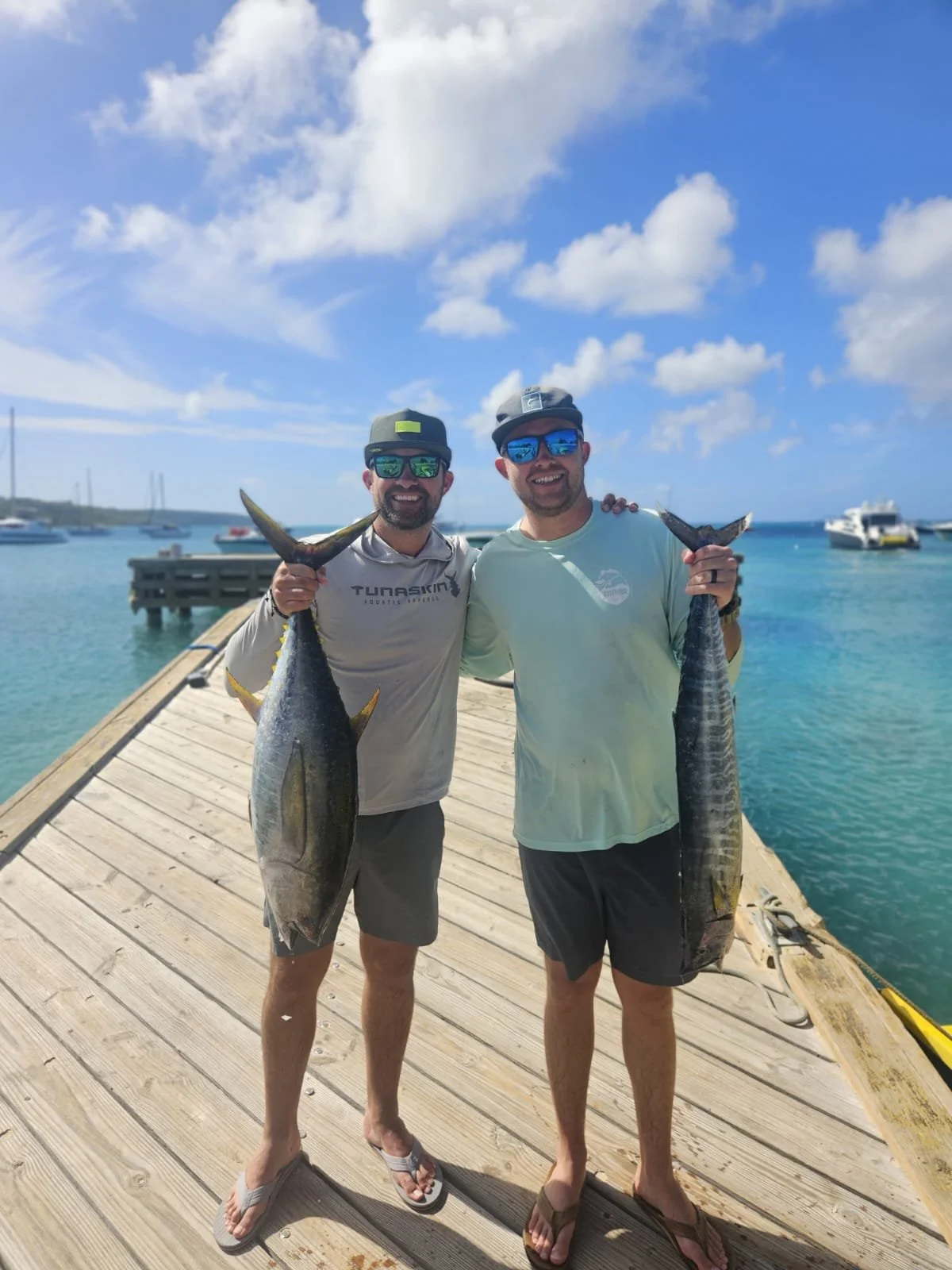 Two men standing on a wooden dock at the water, each holding a large fish they caught, smiling at the camera. They are wearing sunglasses and casual clothing, with boats floating in the background under a blue sky with clouds.