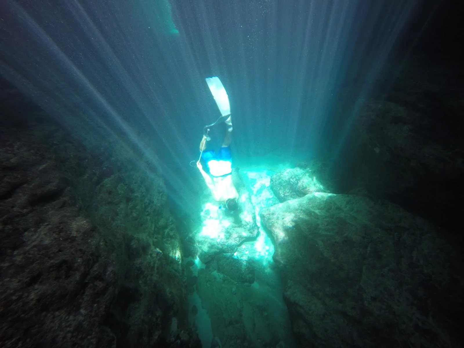 Two scuba divers explore an underwater cave or crevice with sunlight filtering through the water, illuminating the rocky surroundings.