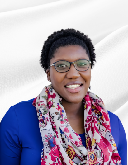 A woman with short curly hair and glasses, smiling, wearing a dark blue top and a colorful scarf, standing against a white wall.