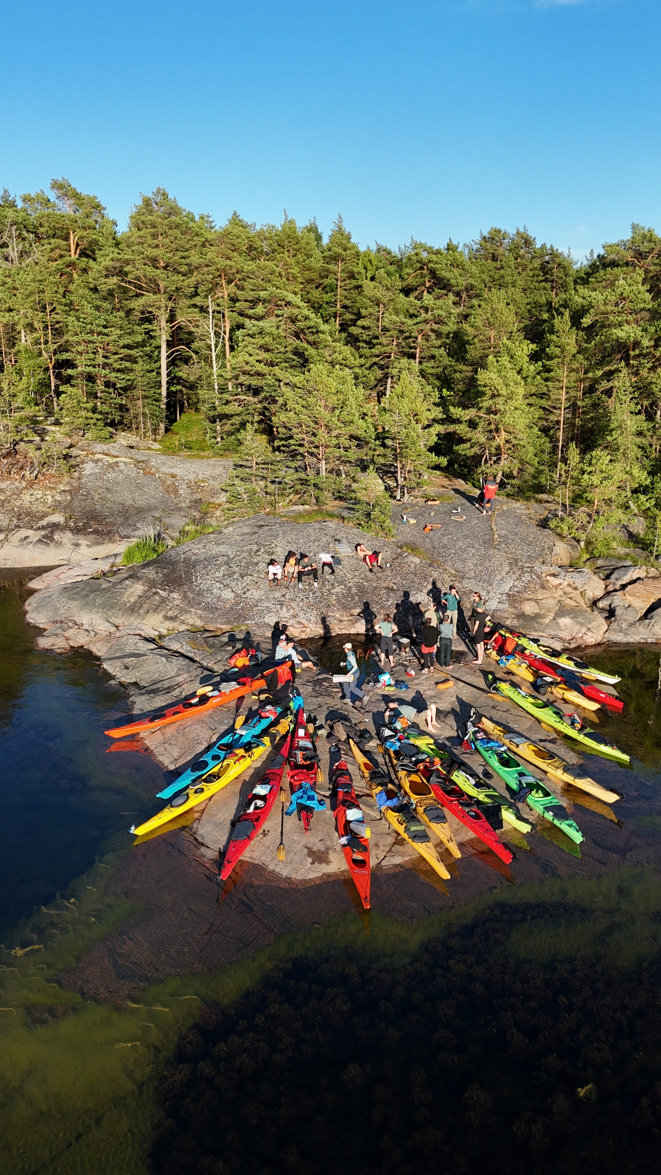 Menschen bereiten sich auf einer Felseninsel mit bunten Kajaks für eine Kanufahrt vor, umgeben von einer Wasserfläche und einem bewaldeten Hintergrund unter einem klaren blauen Himmel.