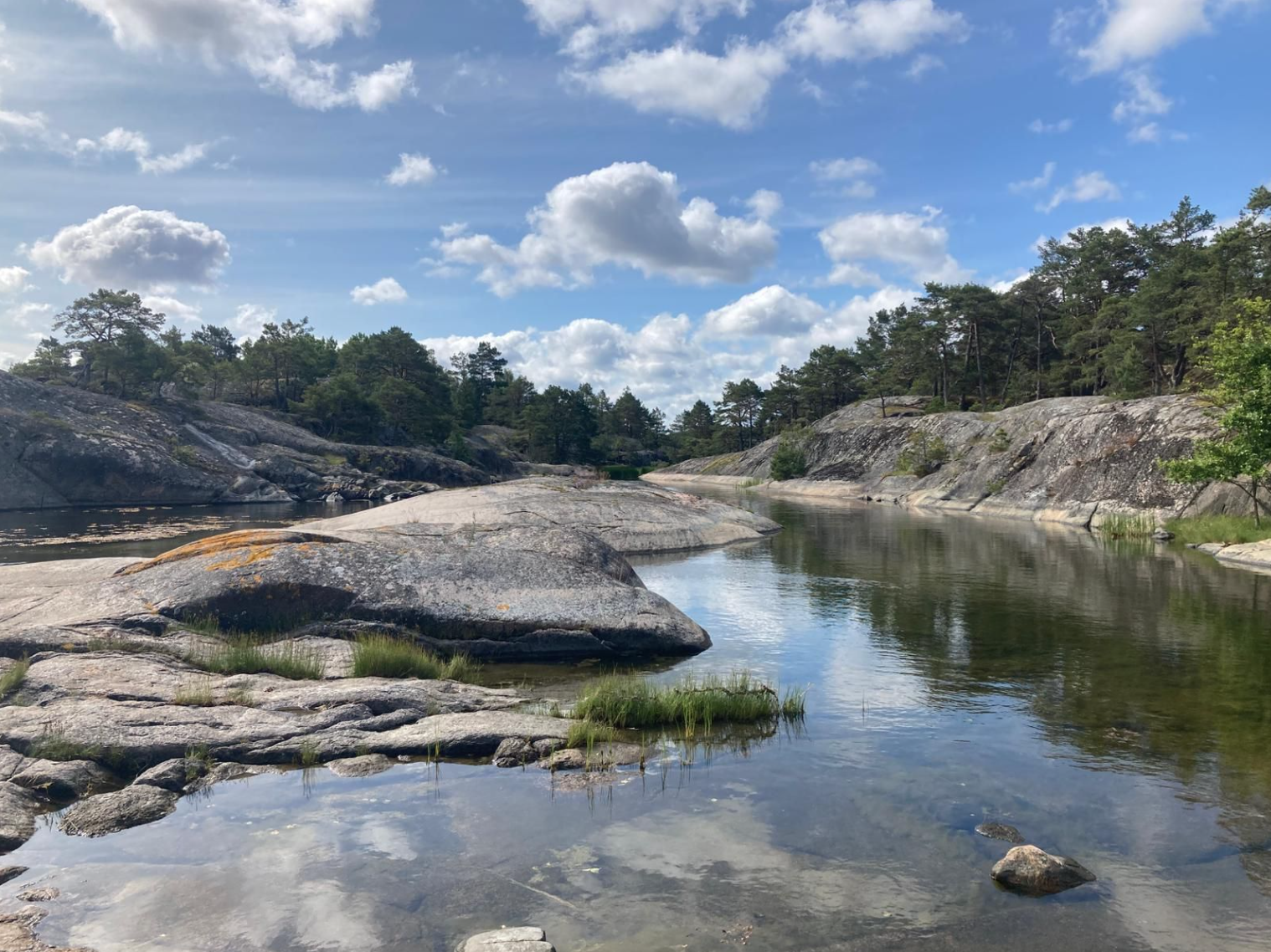 Ein ruhiger Fluss fließt durch eine Landschaft mit großen Felsen und Bäumen, unter einem blauen Himmel mit Wolken.