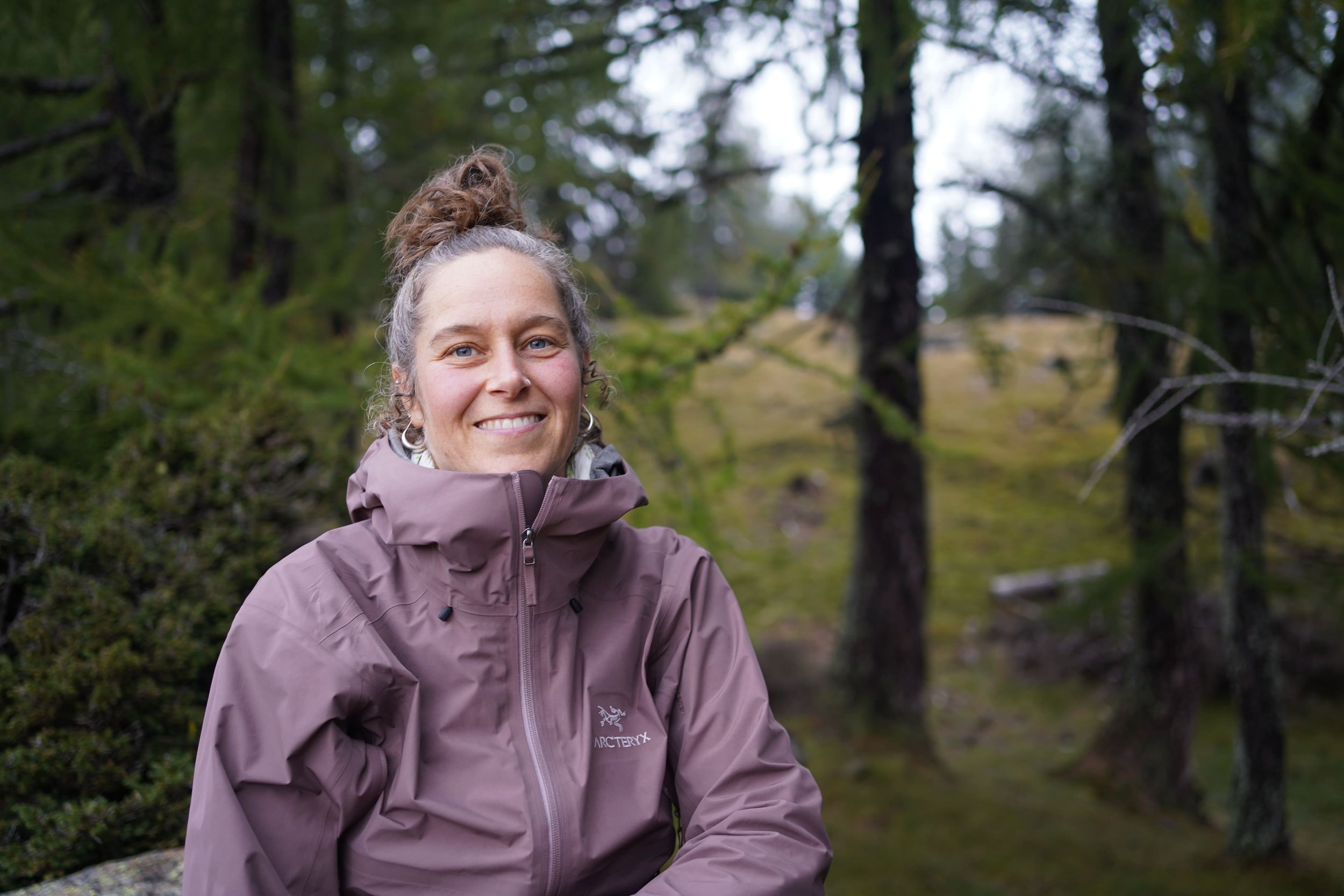 Eine lachende Frau in einer braunen Jacke sitzt im Wald, umgeben von Bäumen und Moos.