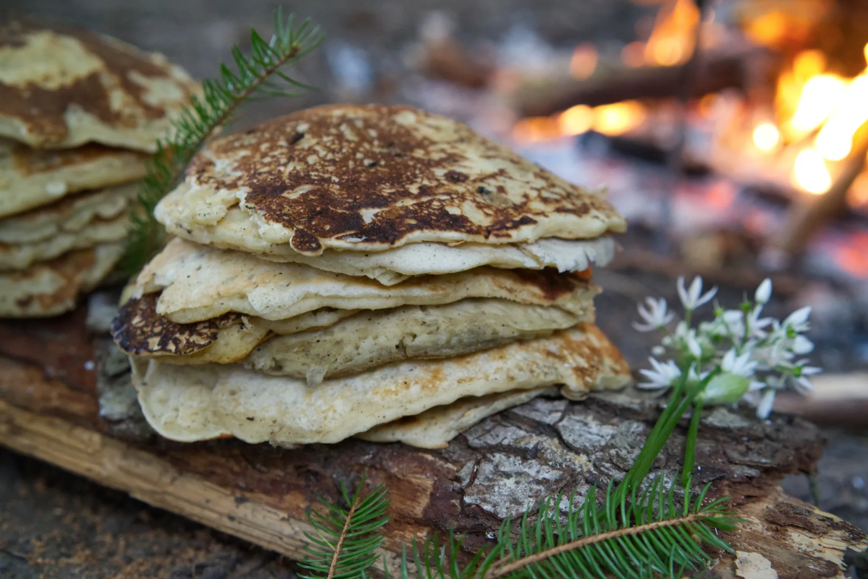 Mehrere Pfannkuchen stapeln sich auf einem Holzstück vor einem Lagerfeuer, mit Tannenzweigen und kleinen weißen Blumen davor.