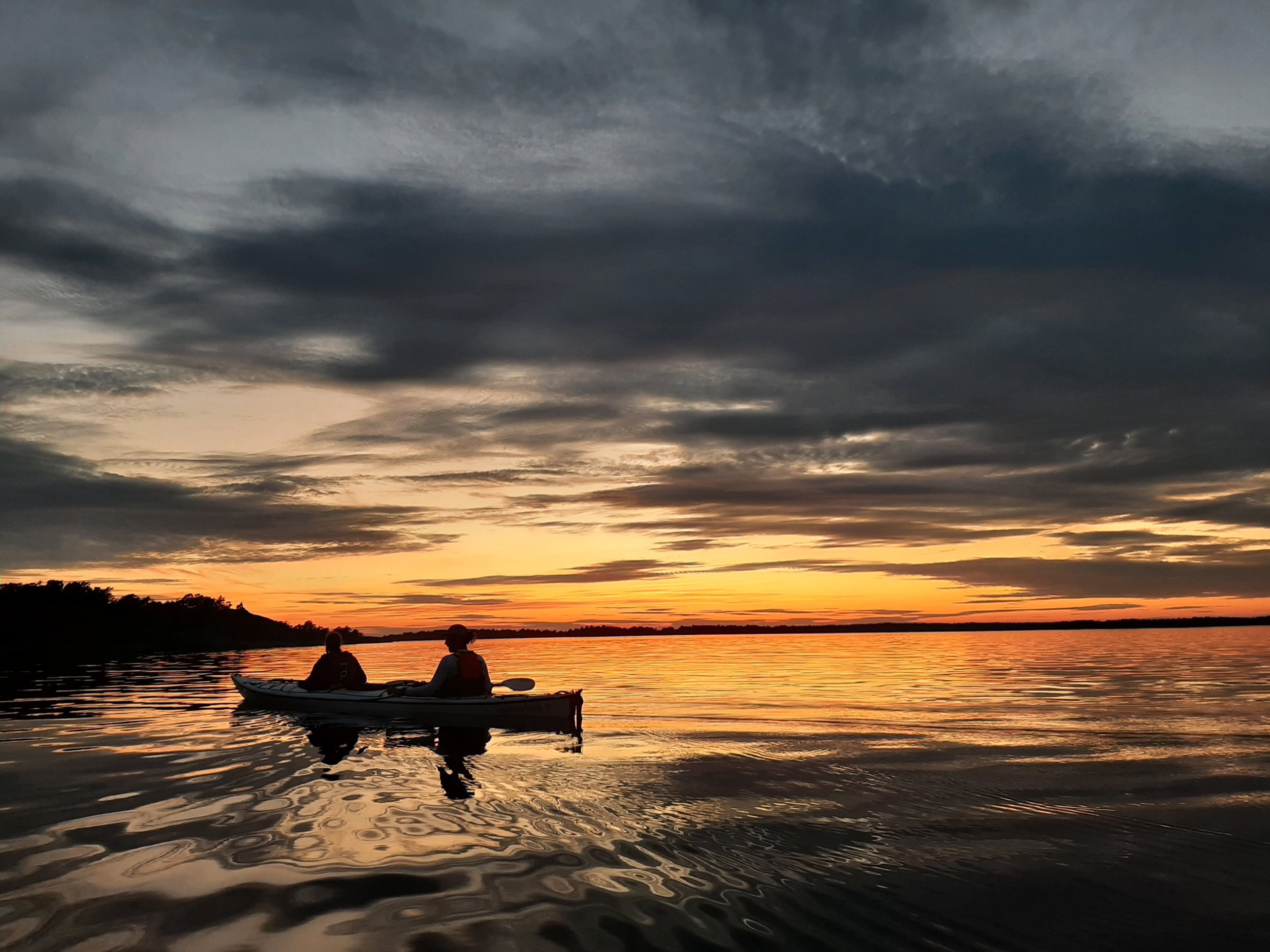 Zwei Menschen in einem Kajak auf einem ruhigen See bei Sonnenuntergang unter einem bewölkten Himmel.