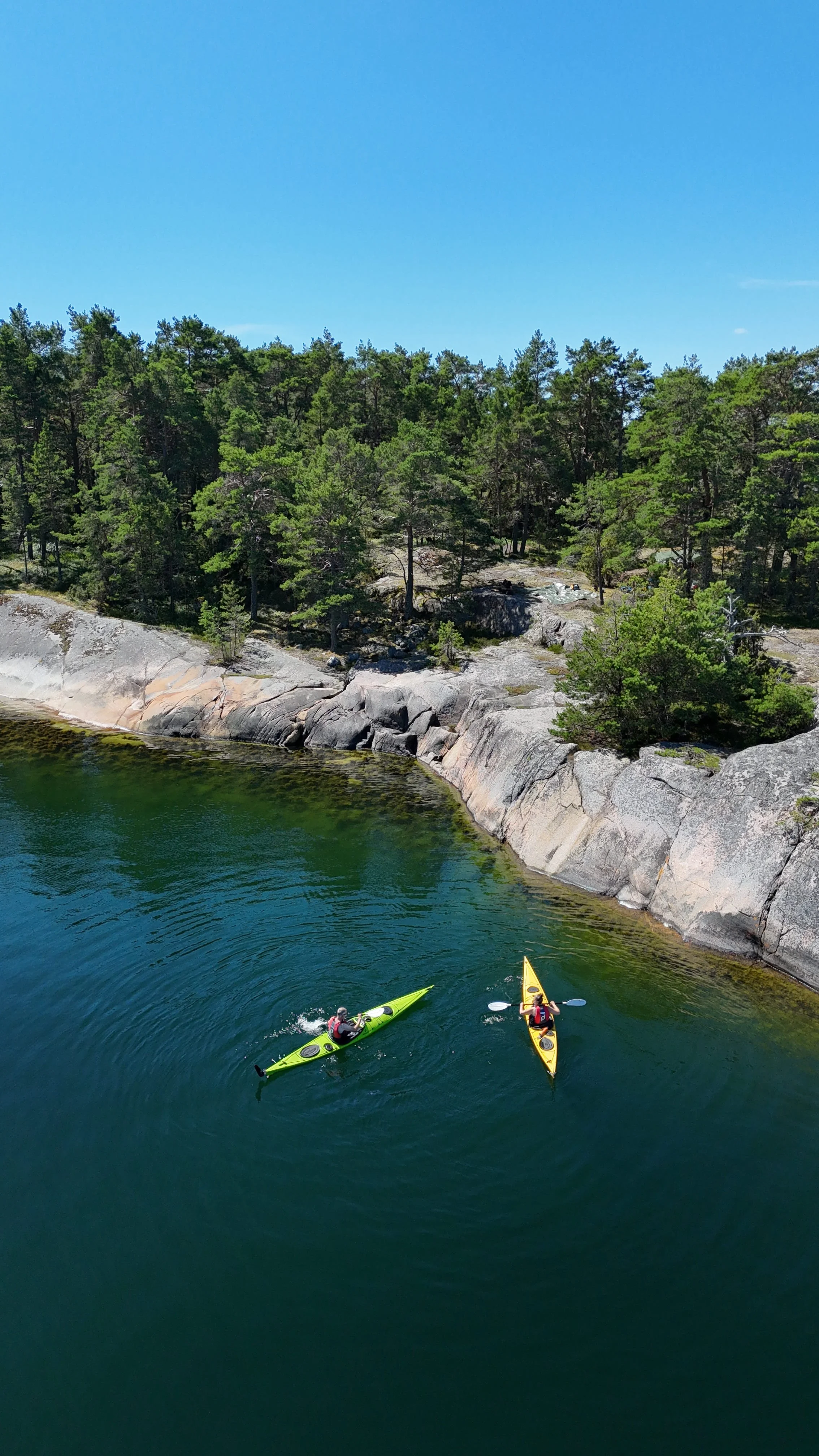 Zwei Personen paddeln in Kajaks auf einem ruhigen Gewässer vor einer Felsenküste mit Bäumen unter blauem Himmel.