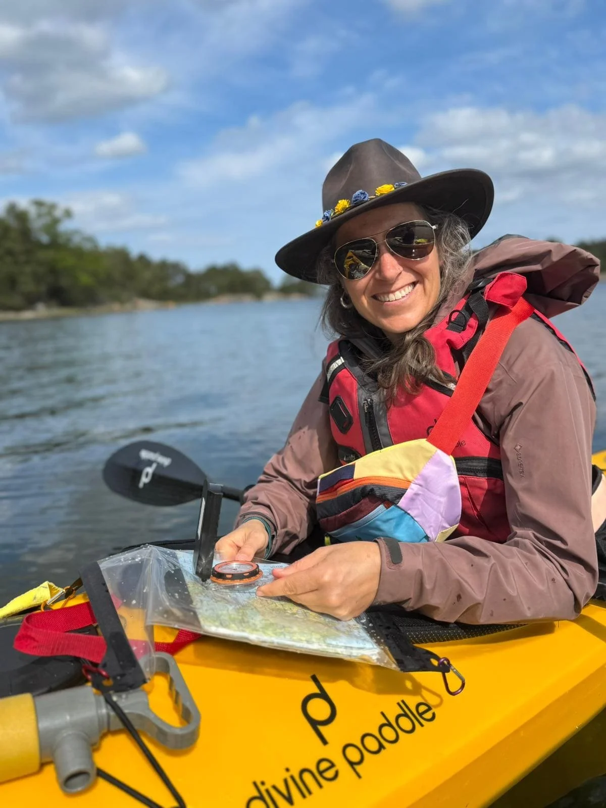 Frau beim Kajakfahren auf einem Fluss, tragt Sonnenbrille und Hut, lächelt, hält eine Wasserkarte, umgeben von Natur, am Himmel sind Wolken.