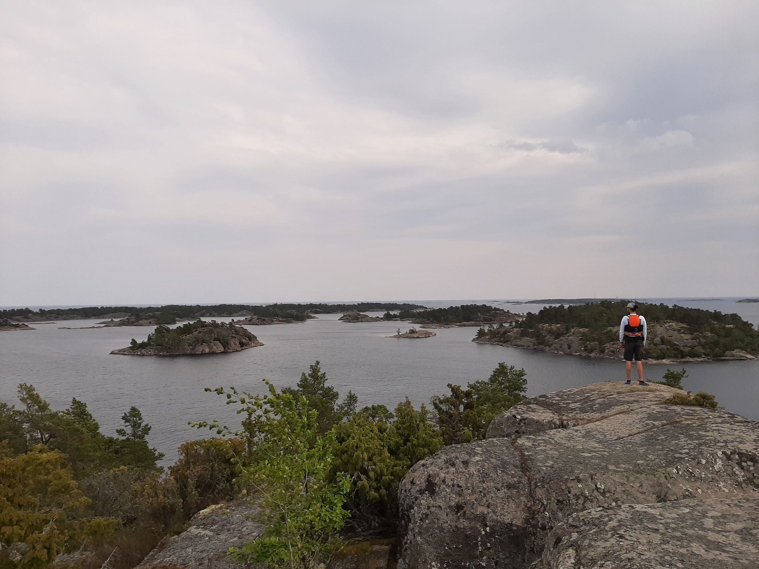 Ein Mann mit Schwimmweste steht auf einem Felsen und blickt auf eine Inselwelt mit kleinen Inseln im Wasser unter einem bewölkten Himmel.