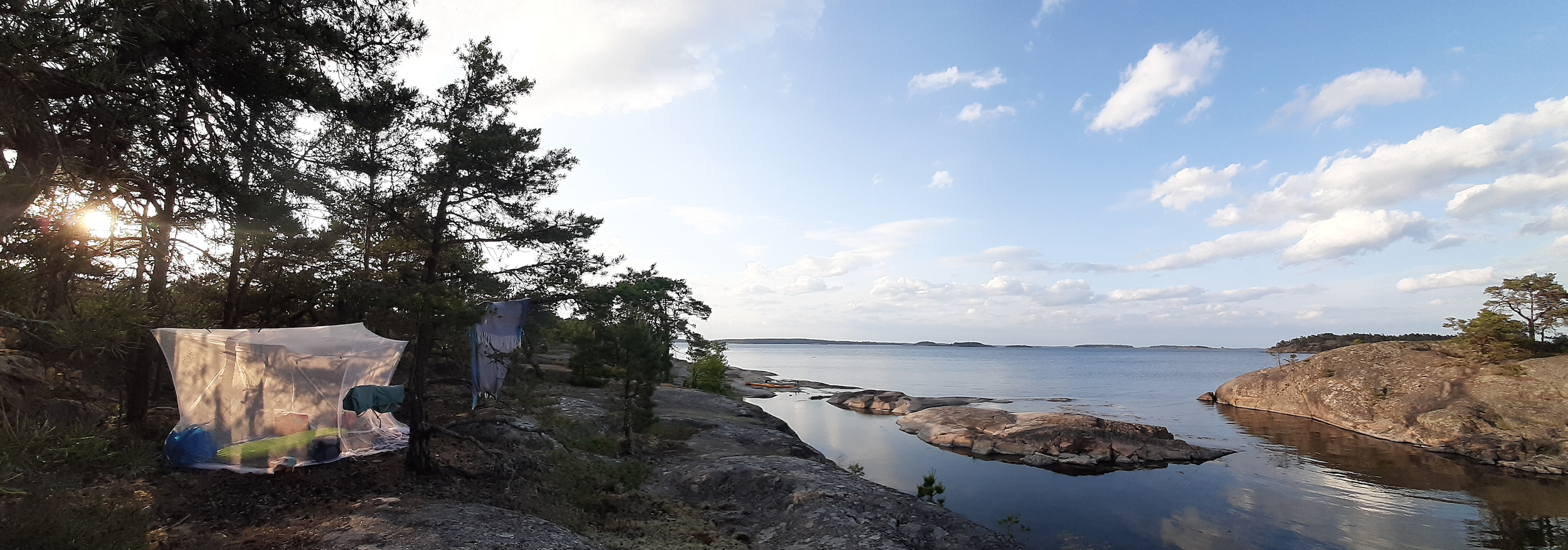 Das Meer mit Felsen und Bäumen, das Wasser spiegelt den Himmel mit Wolken wider. Auf dem linken Bildrand ist ein feuchtes Tuch, vermutlich ein Zelt, bei Sonnenuntergang.