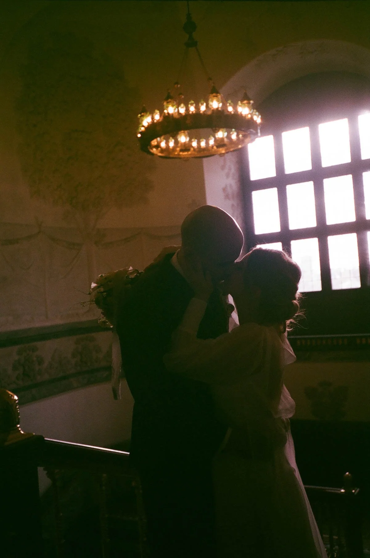 A silhouette of a bride and groom sharing a kiss inside a church with a large window and a chandelier hanging from the ceiling.